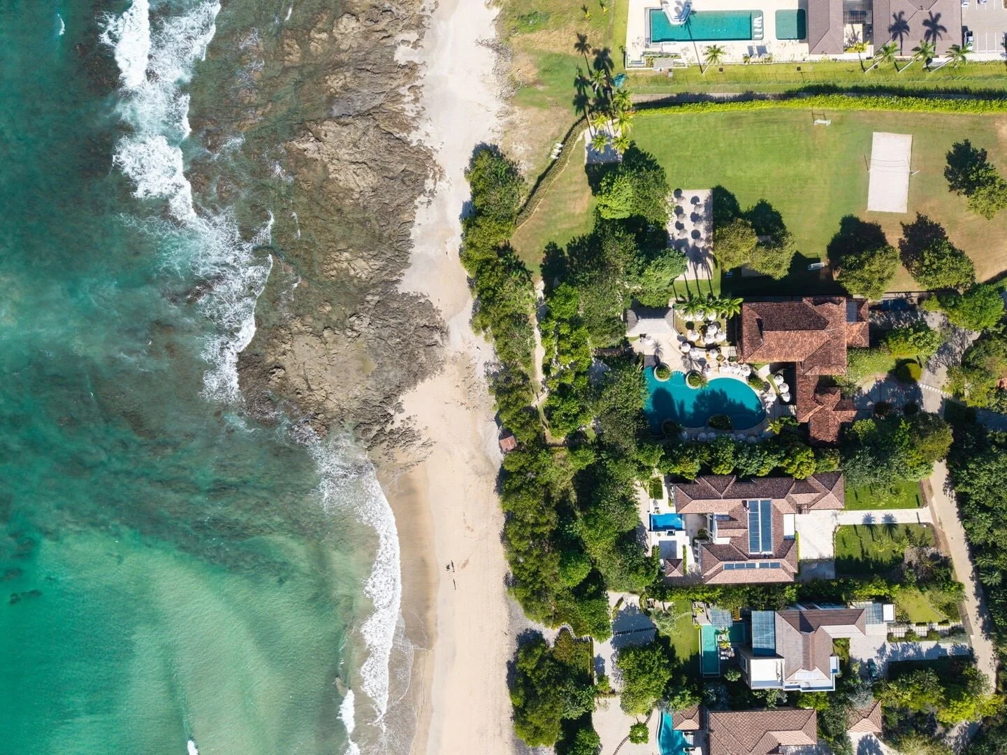 Aerial view of a beachside residential area with houses, a swimming pool, lush green lawns, and a sandy beach with waves hitting the shore.