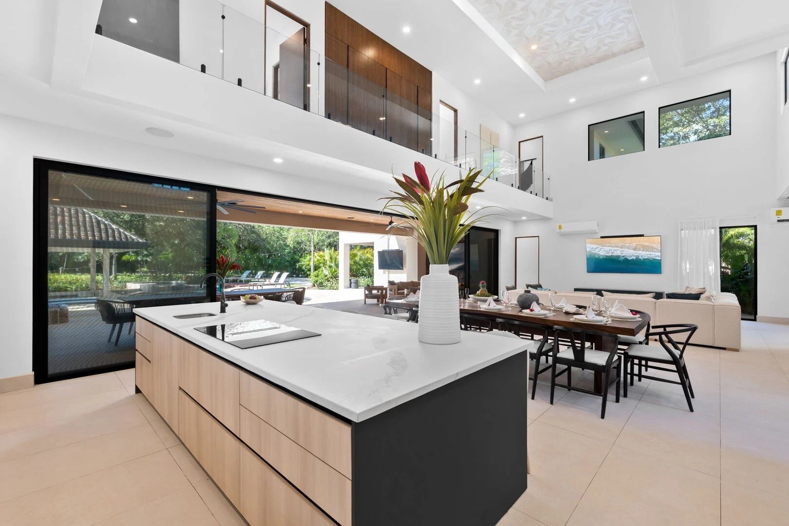 Modern open-concept living space with a kitchen island, dining area, and living room, featuring large windows and a balcony, with outdoor view including a pool and greenery.