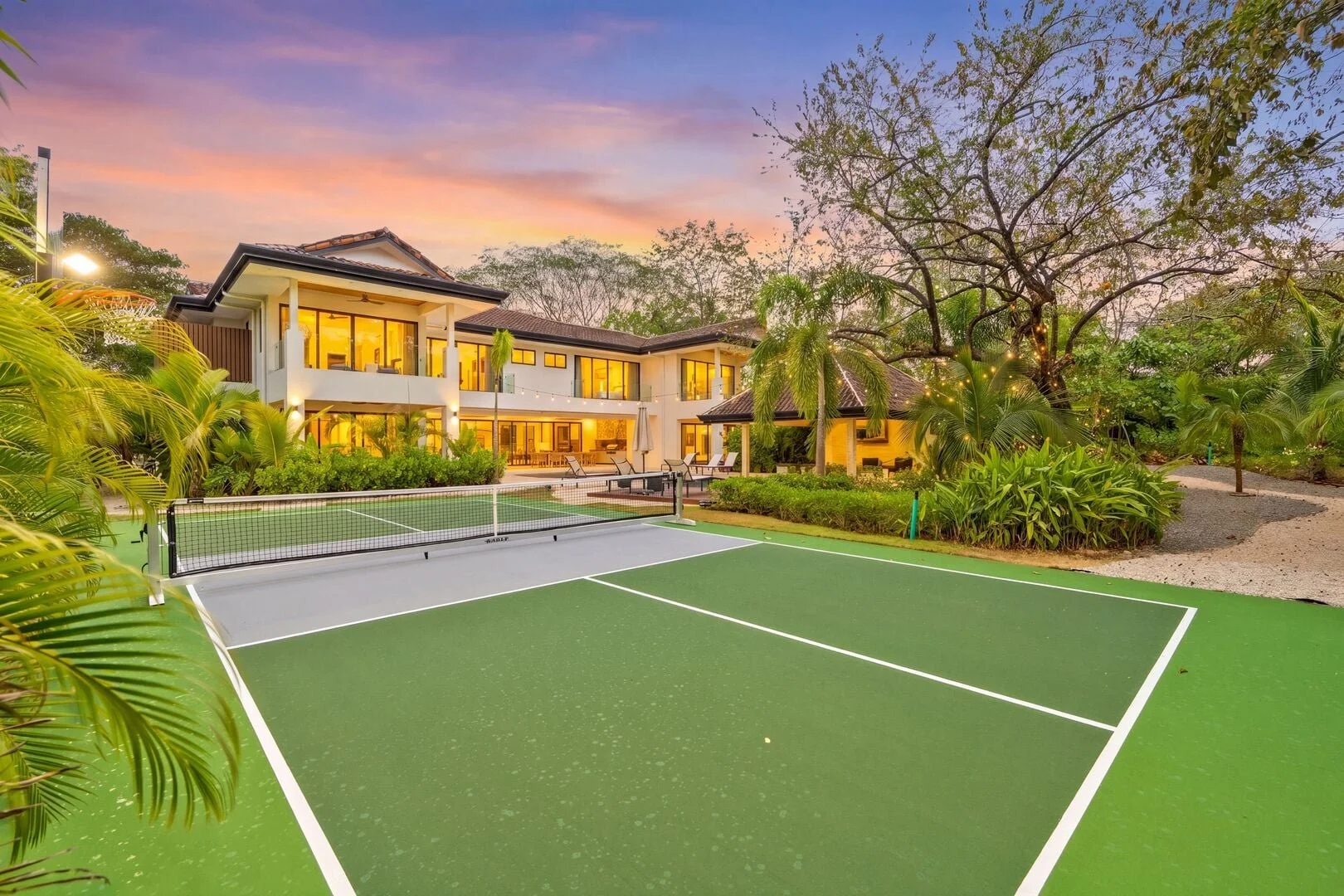 Luxury house with lit windows in evening, surrounded by lush greenery, with a tennis court in the foreground.