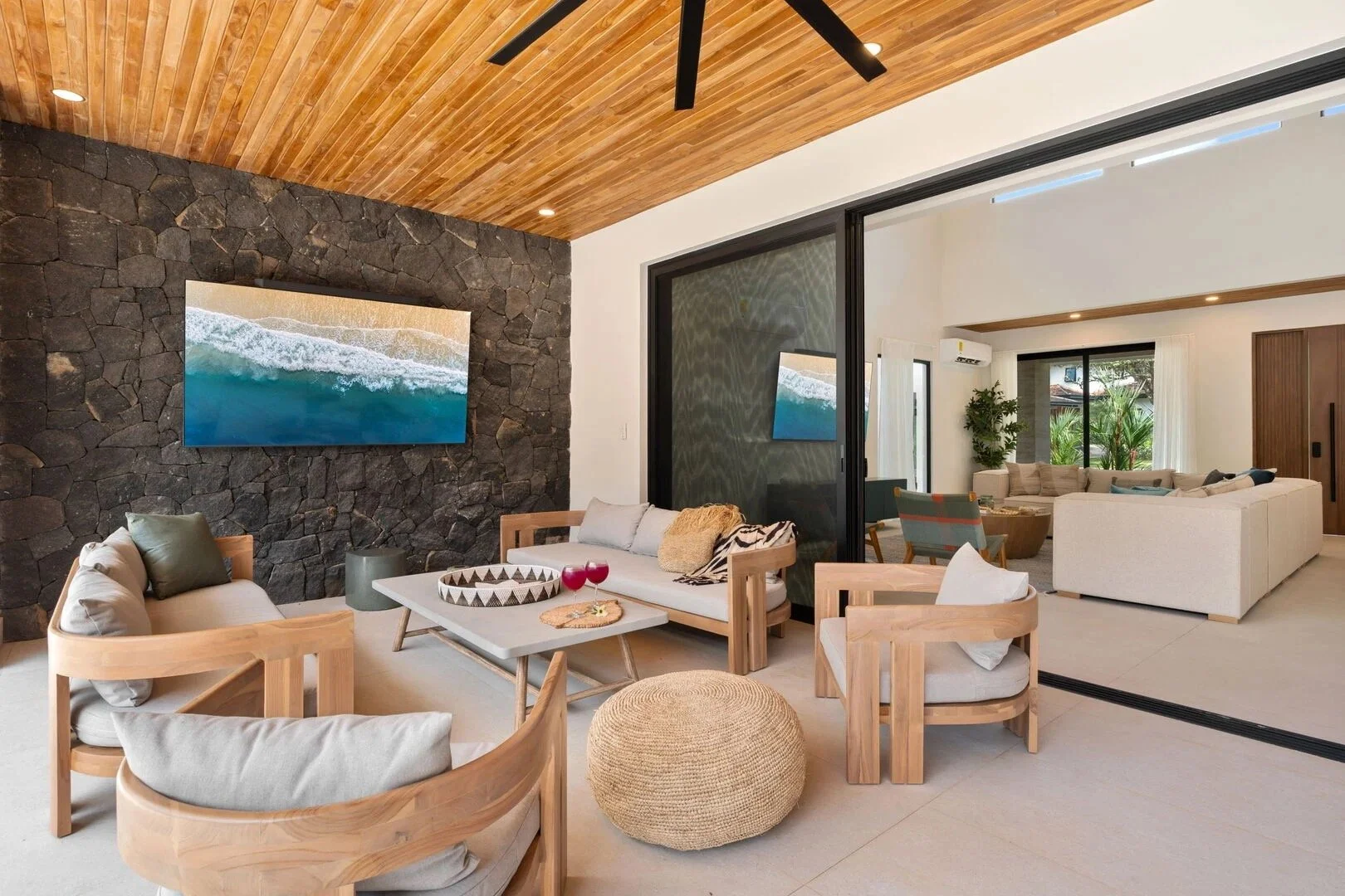Modern living room with stone accent wall, wooden ceiling, and large white sectional sofa; TV on wall, glass sliding door, and plants in background.