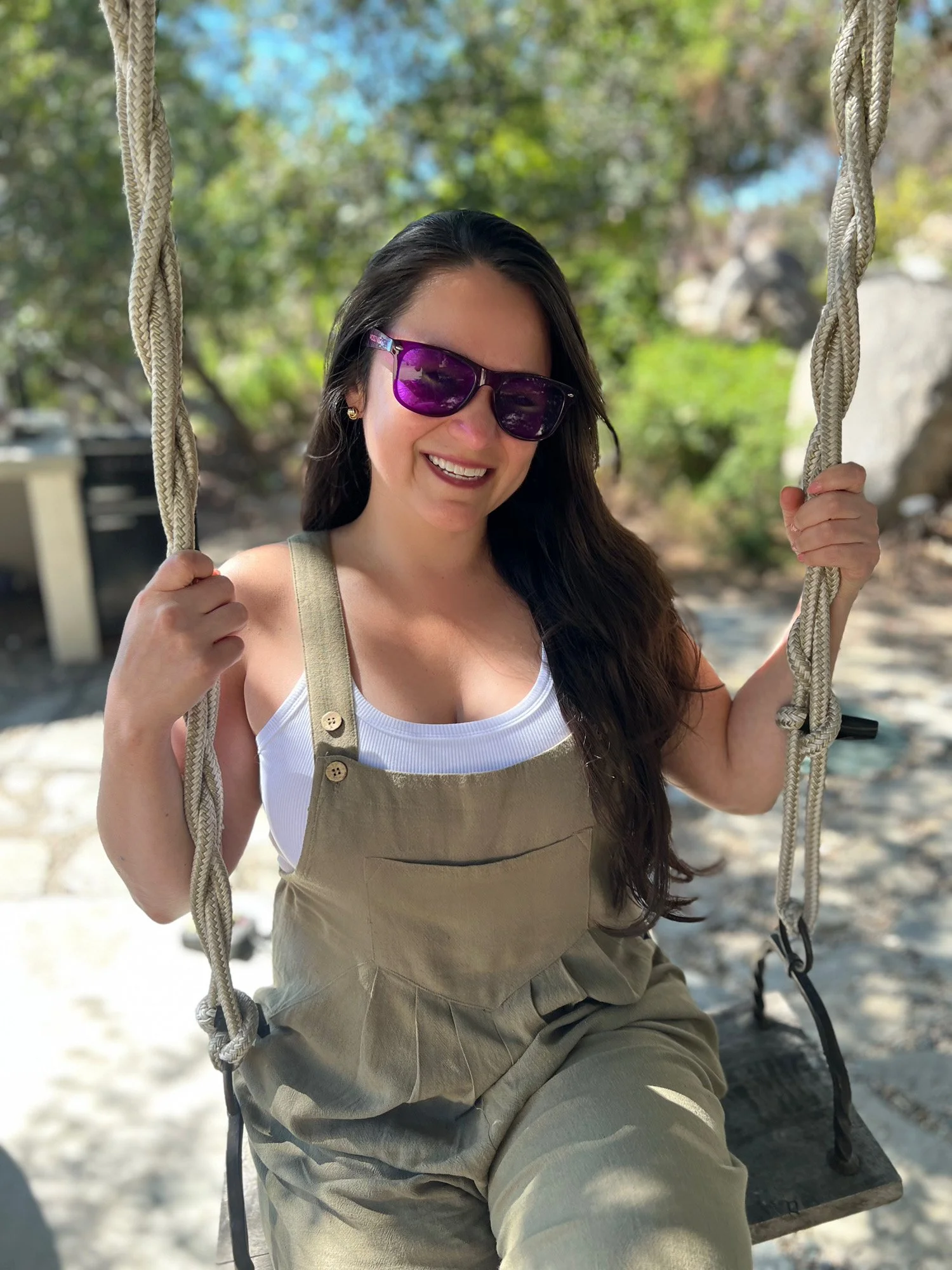 A smiling woman with long dark hair, wearing purple sunglasses, a white tank top, and khaki overalls, sitting on a swing outdoors with green trees and rocks in the background.