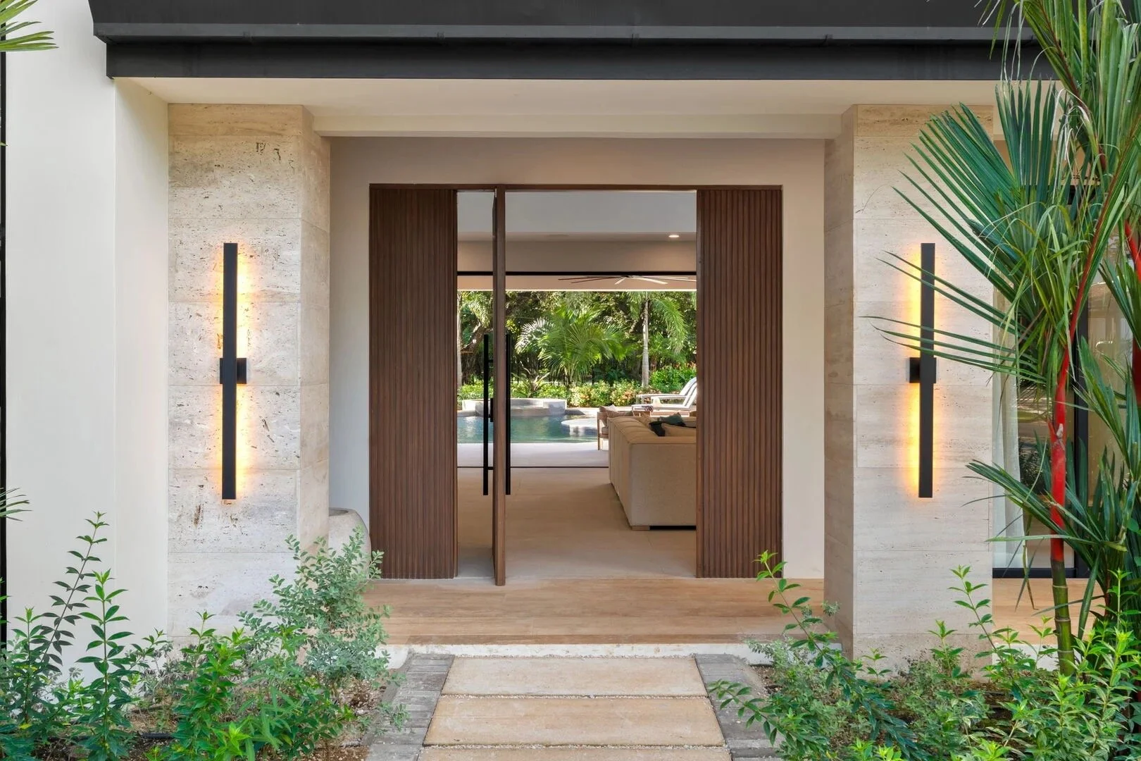 Modern house entry with wooden sliding doors, stone walls, outdoor plants, and a view of a backyard with a pool and lush greenery.