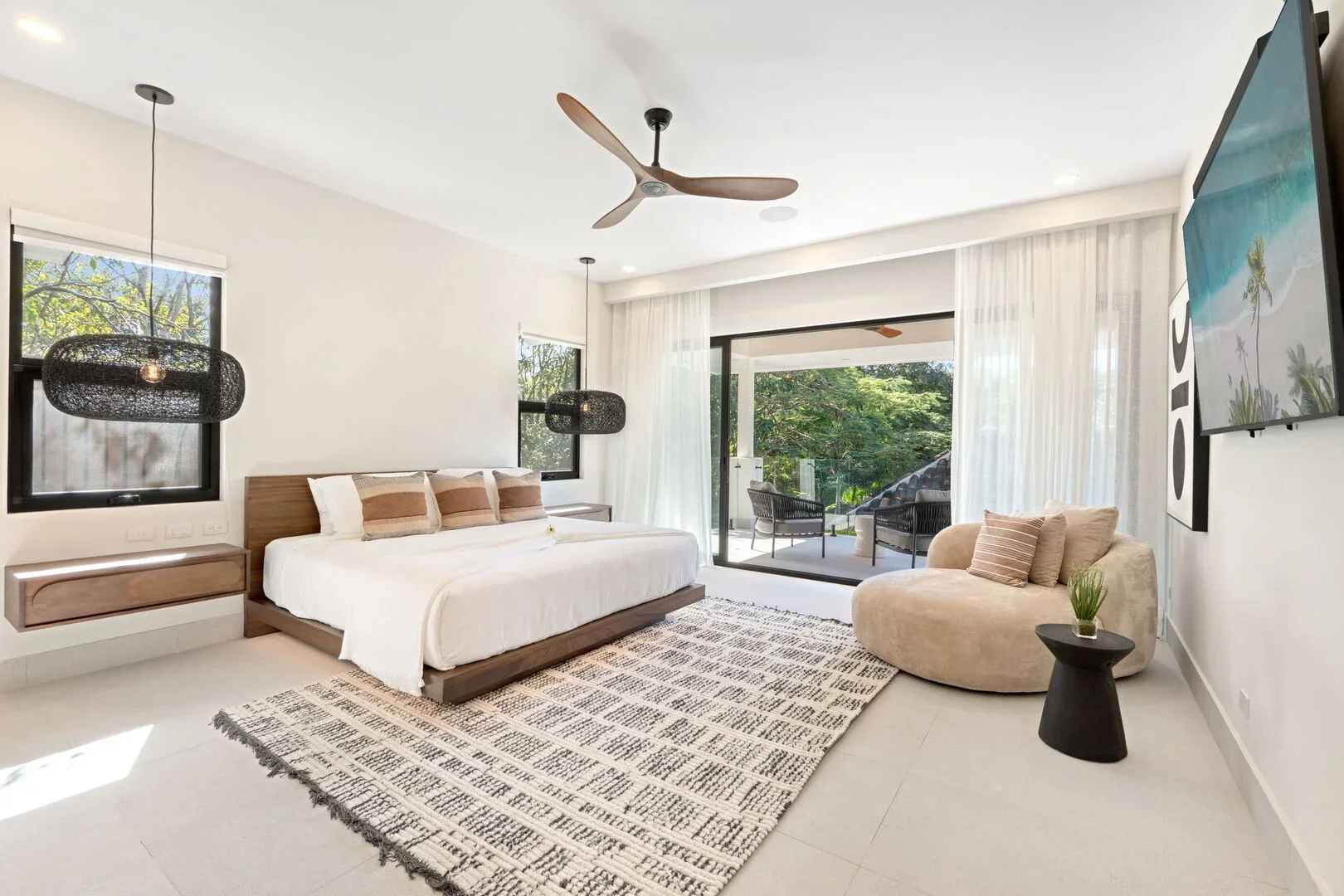 A modern bedroom with a large bed, two black pendant lights, a beige sofa with pillows, and a sliding glass door leading to an outdoor patio with chairs and trees visible through the windows.