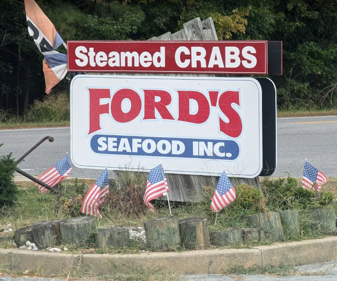 A roadside sign for Ford's Seafood Inc., advertising steamed crabs, decorated with small American flags.
