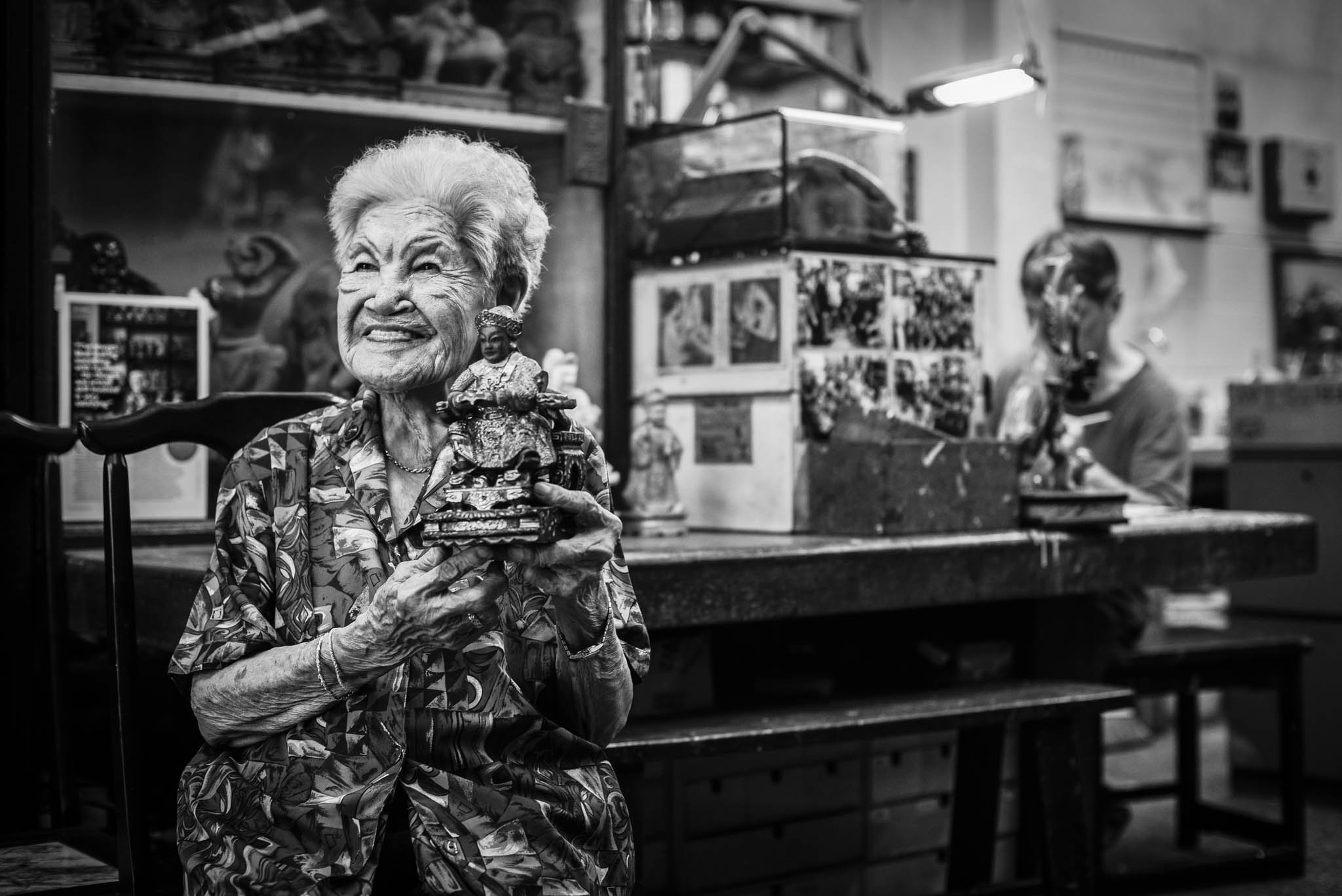 Mdm Tan poses for a portrait with a finished effigy, while her son Ng Yeow Hua puts finishing touches to other effigies at his work desk.