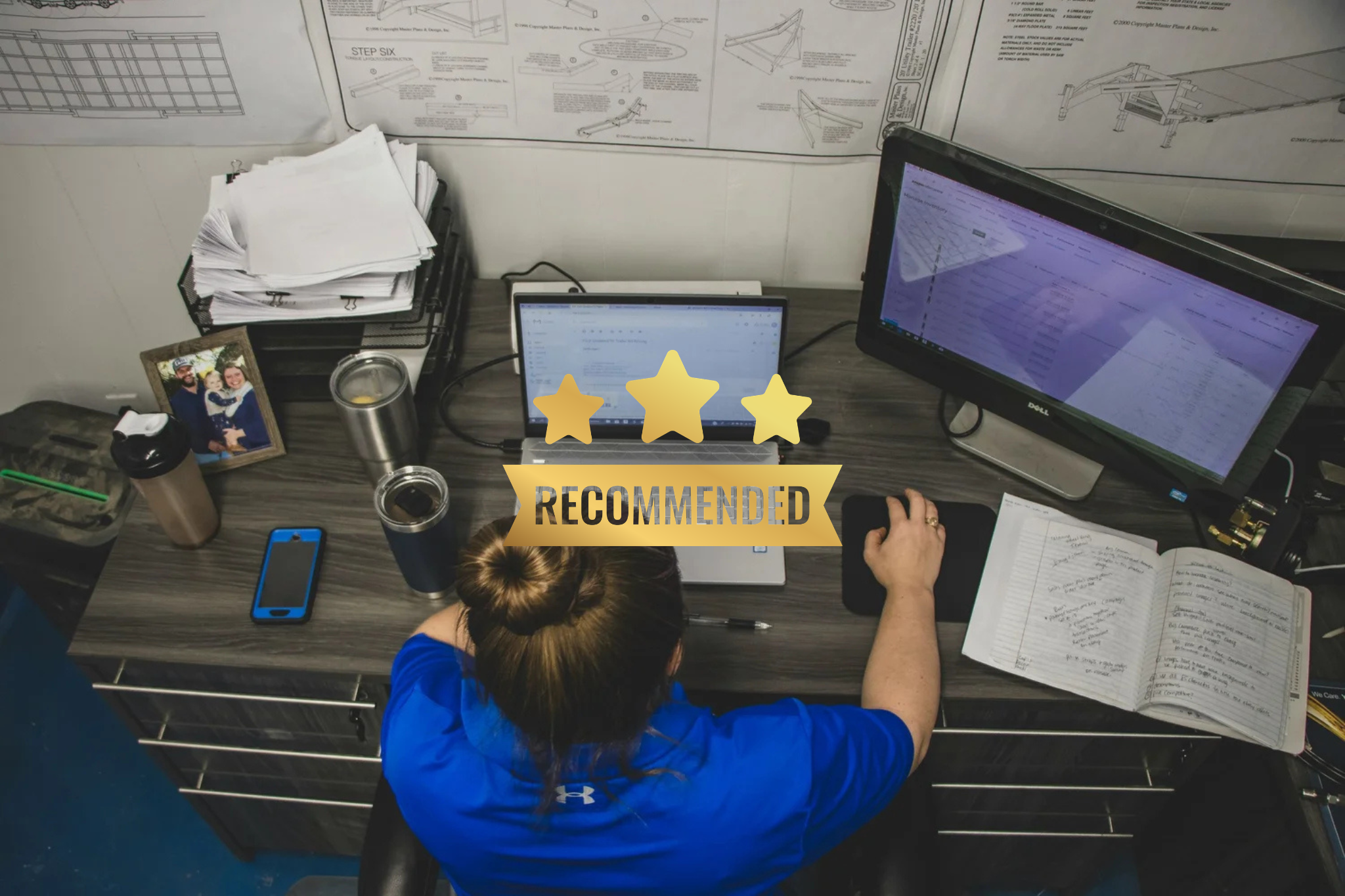 Top-down view of a cluttered desk with a woman working, multiple documents, two monitors, a smartphone, notebooks, a photograph, and various office items, with a 'Recommended' badge overlay.