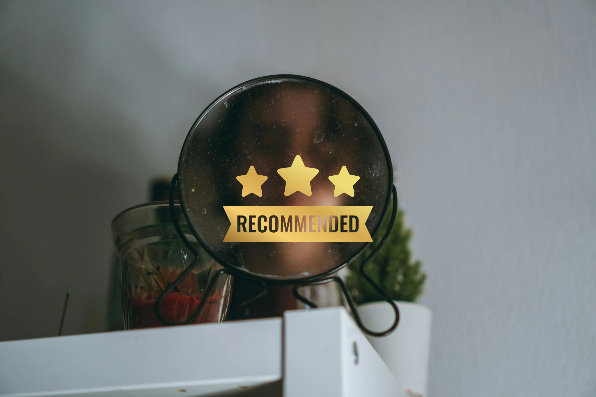 A person taking a photo of themselves using a small mirror with a sticker that has three gold stars and a banner reading 'RECOMMENDED' on it, with a potted plant and a glass container nearby.