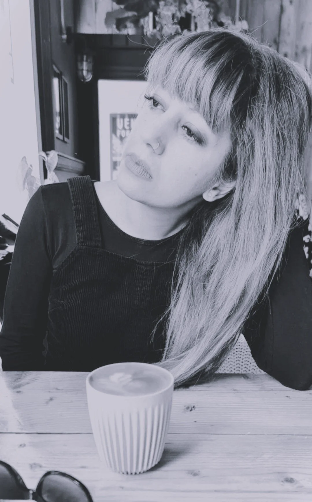 Black and white photo of a woman with long hair, sitting at a table, looking thoughtfully to the side, with a cup of latte and sunglasses on the table.