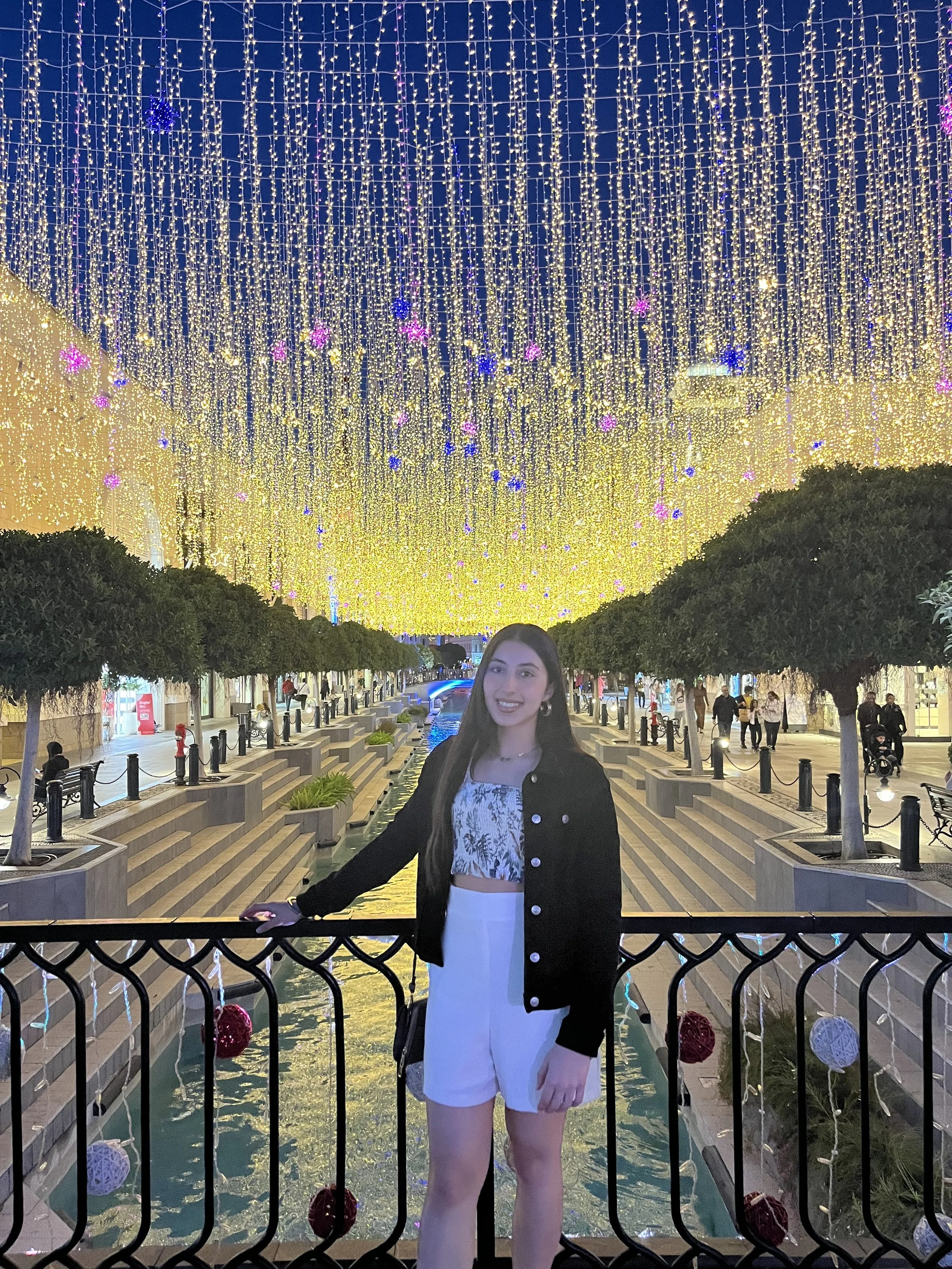 A young woman with long dark hair and a big smile stands on a bridge with a black railing, in front of a canal lined with trees. Above, an elaborate display of multicolored string lights decorates the sky, creating a festive atmosphere at night.