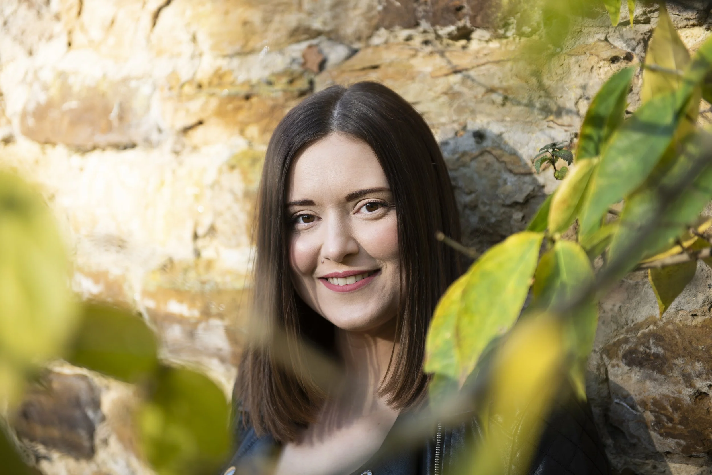 A young woman with long brown hair and light skin smiling at the camera, partially obscured by green leaves, standing against a rough stone wall.