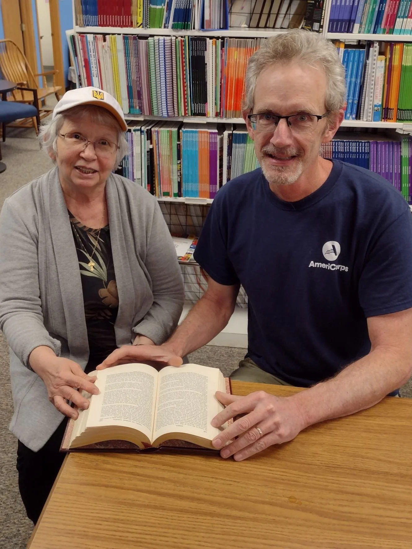 Man and woman sitting next to each other holding open a book on the table in front of them. They are sitting in front of a large bookshelf full of colorful books.