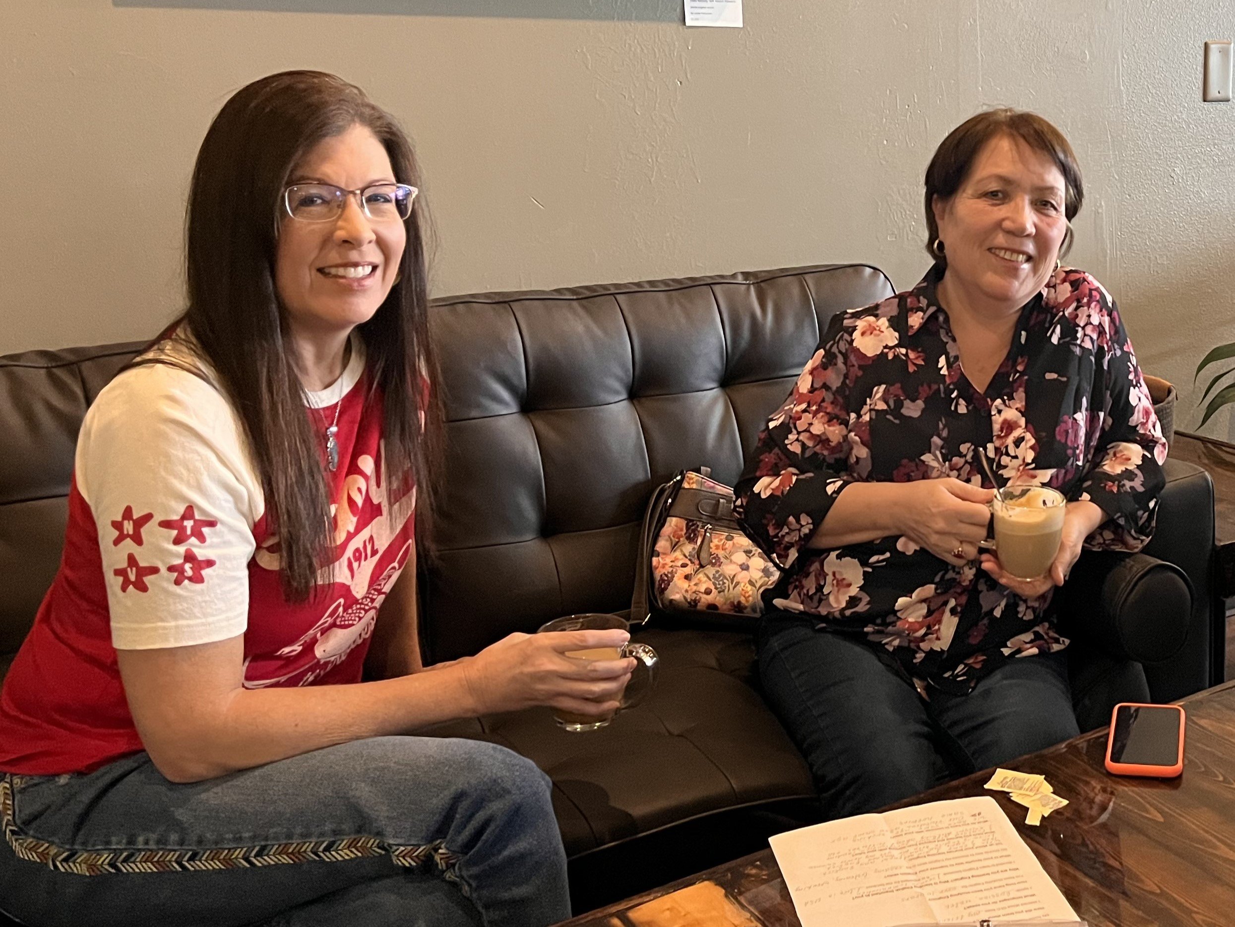Two white women sitting on a leather couch drinking coffee together