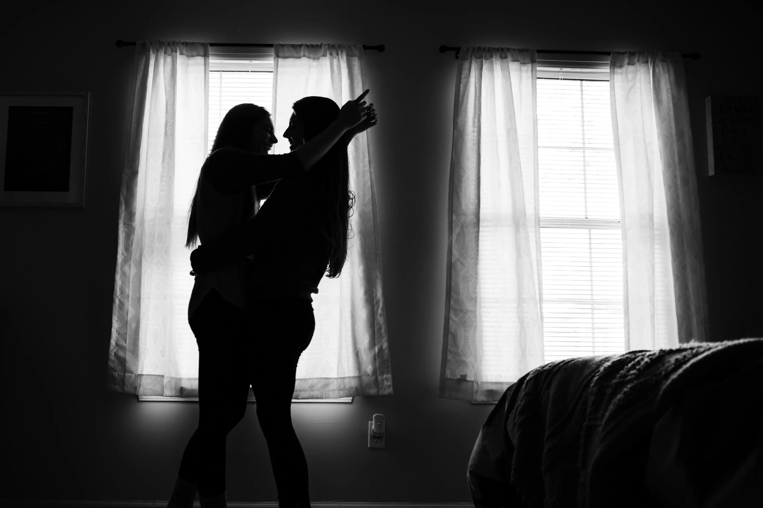 Two women are embracing and smiling in front of a window with sheer curtains, backlit by natural light in a bedroom, representing online couples therapy.