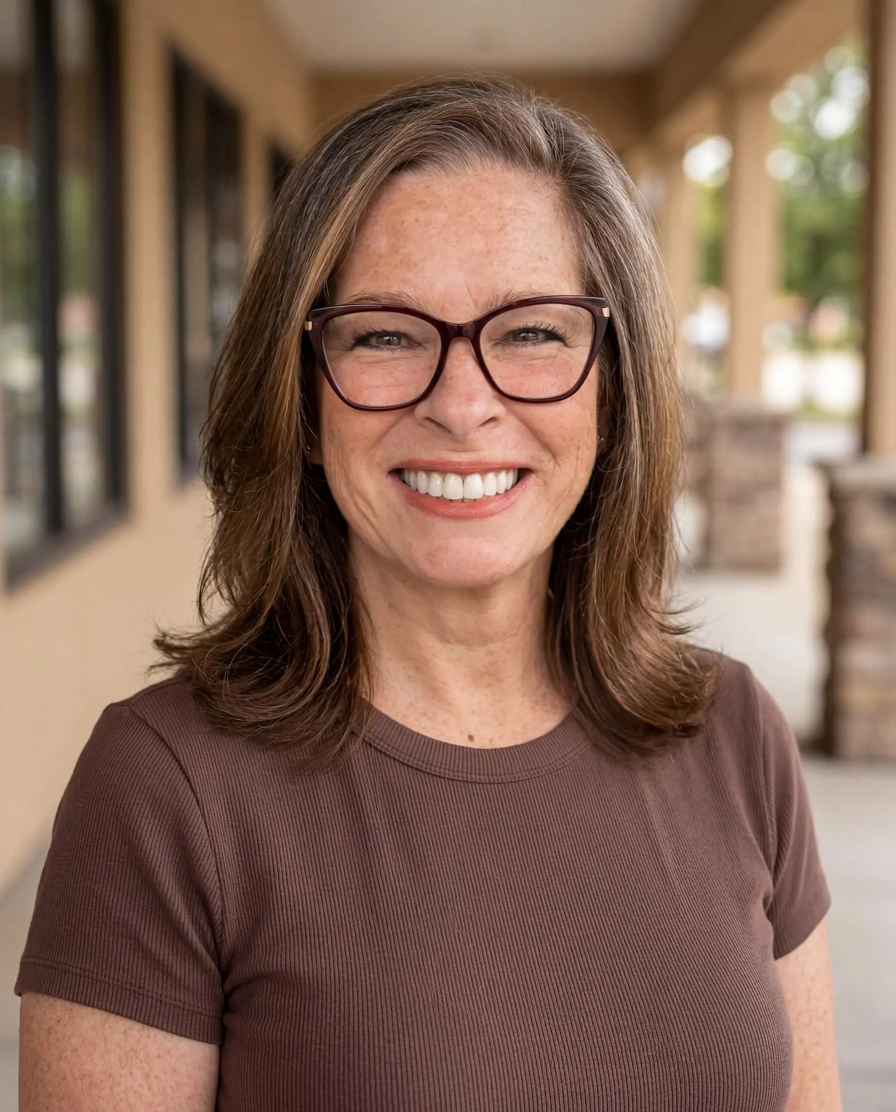 A smiling woman (online sex therapist Jan Carey) with long brown hair wearing glasses and a brown shirt, standing outdoors with a blurred background of windows and greenery.