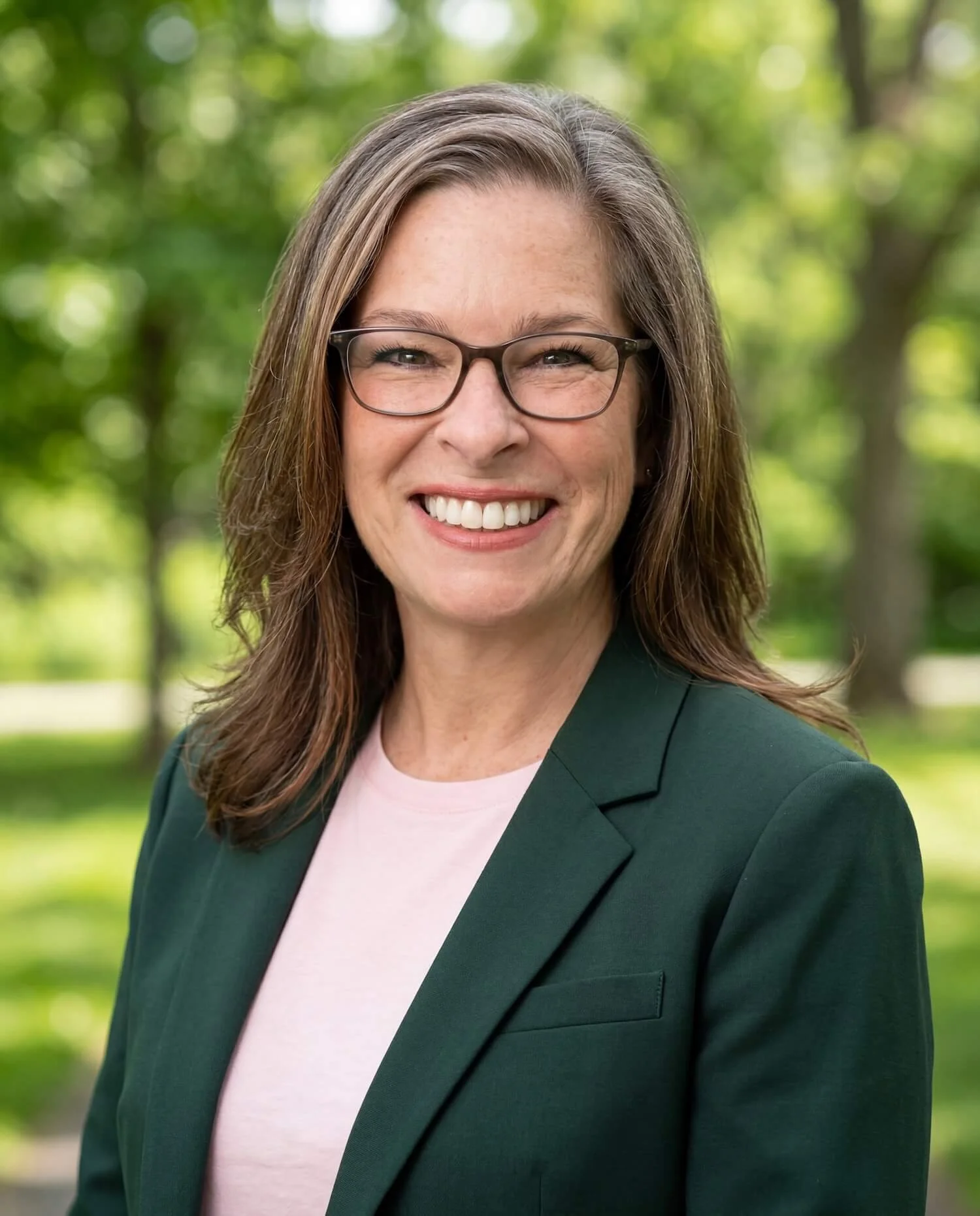 Online couples therapist Jan Carey, a woman with brown hair, glasses, and a bright smile, wearing a dark green blazer and a light pink shirt, standing outdoors with green trees in the background.