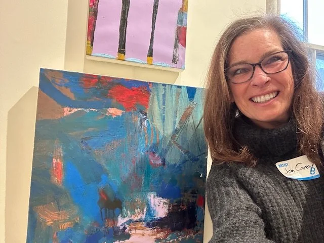 Online couples counselor Jan Carey, awoman with glasses and long brown hair smiling, standing next to an abstract painting with blue, red, pink, and black colors.