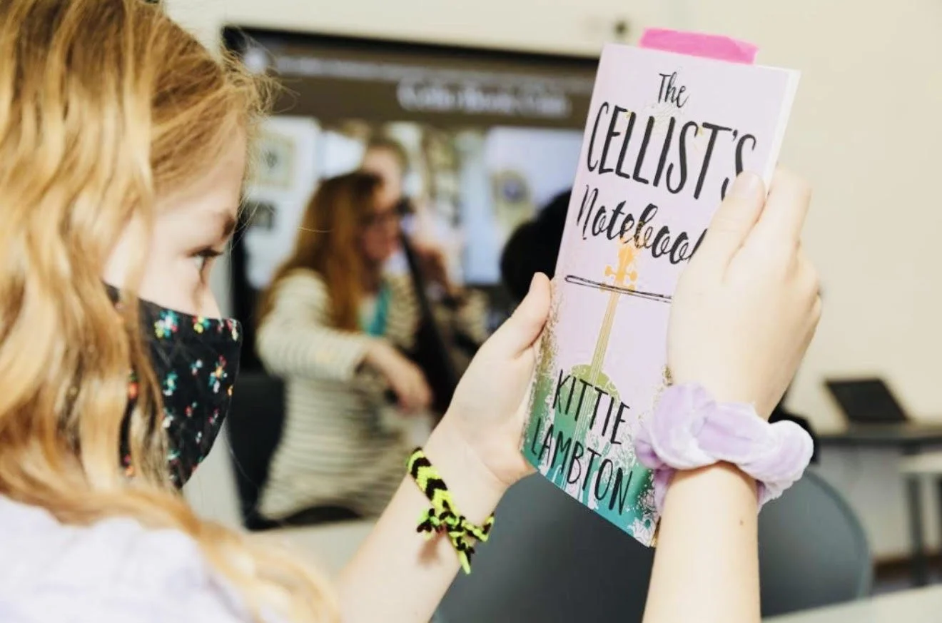 A young woman with red hair, wearing a black face mask with colorful dots, holds a book titled 'The Cellist's Notebook' by Kittie Lambo. She is seated indoors, with other people in the background.
