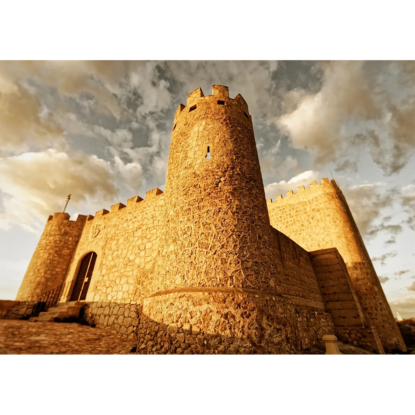 Low-angle fine art print of the 15th-century Spanish Castillo de Jumilla bathed in warm golden hour light against a swirling sky.