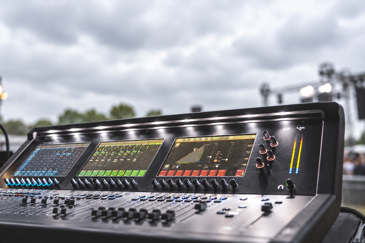A digital sound mixing console with multiple screens, knobs, and sliders, set outdoors under a cloudy sky with stage lighting equipment in the background.