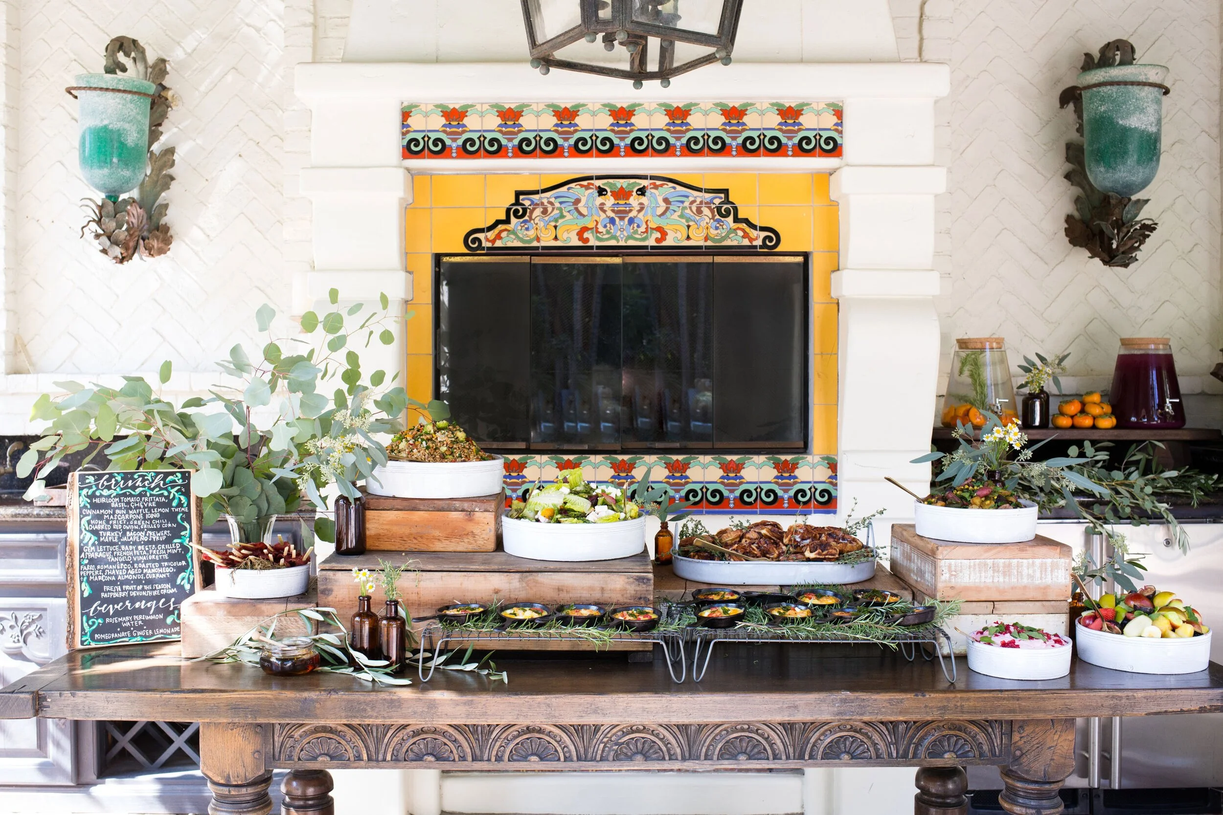 Decorative Buffet style tablescape on a vintage style wooden table top with eucalypt floral arrangements sitting in front of a colorful, yellow tiled fireplace