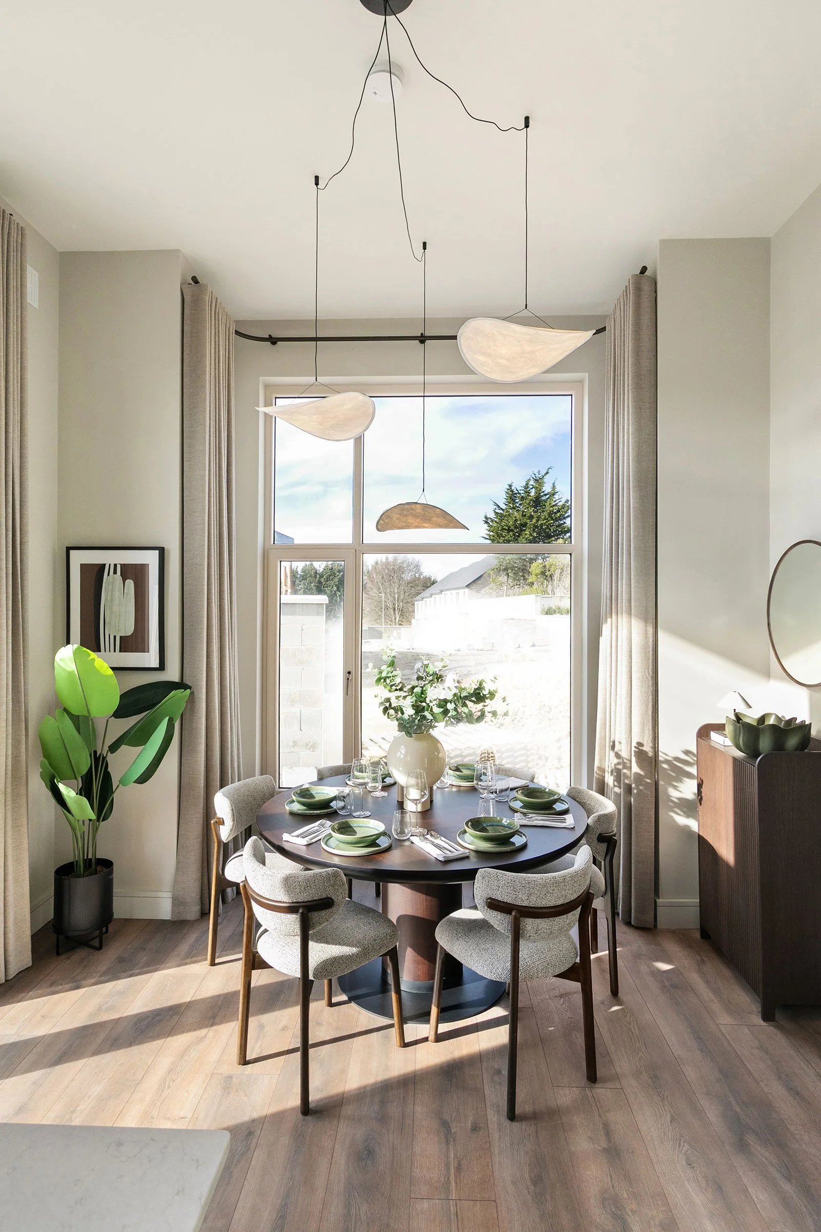 Round dining table with 4 chairs placed in front of large window, striking light fixture and plants