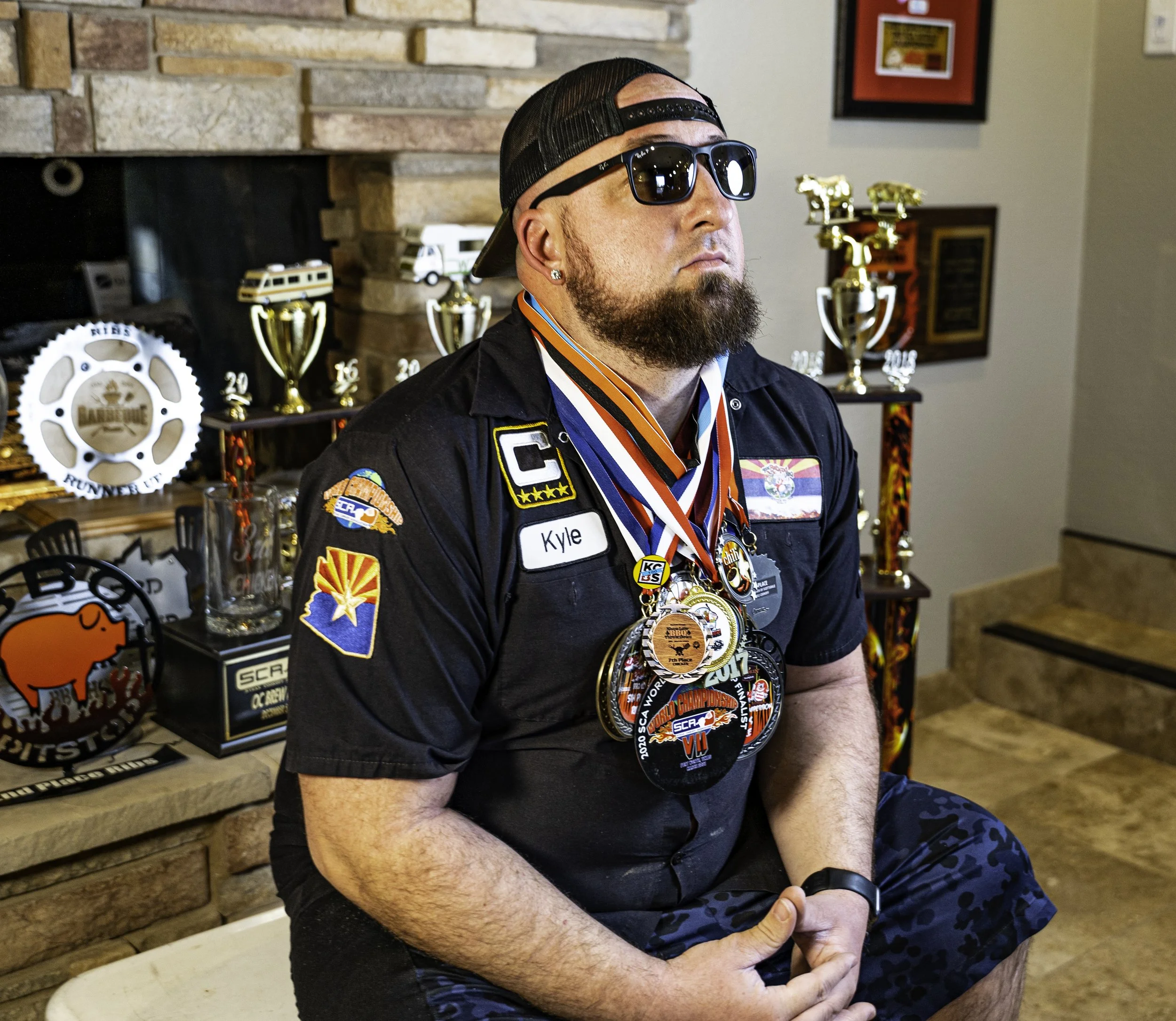 A man with sunglasses, a backwards cap, and a beard, wearing multiple medals around his neck, sitting on a bench inside a room with trophies and framed pictures in the background.