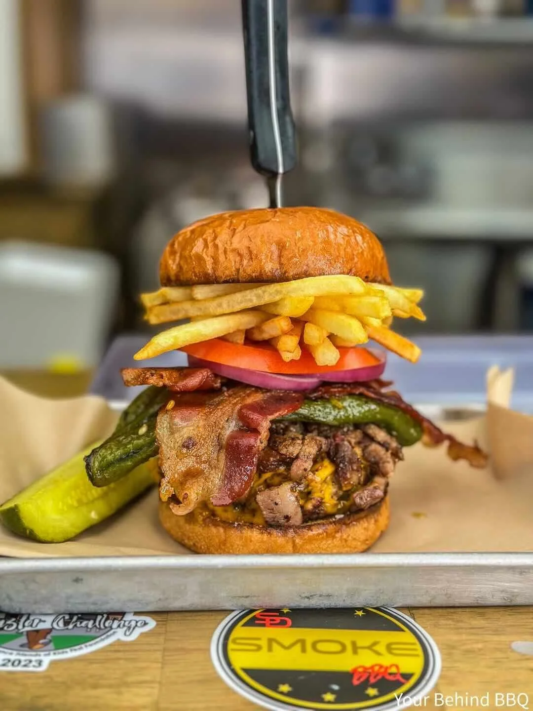A towering burger with a bun, cheese, grilled beef patty, bacon, pickles, lettuce, tomato, onions, French fries, and a knife stabbed through the top bun.