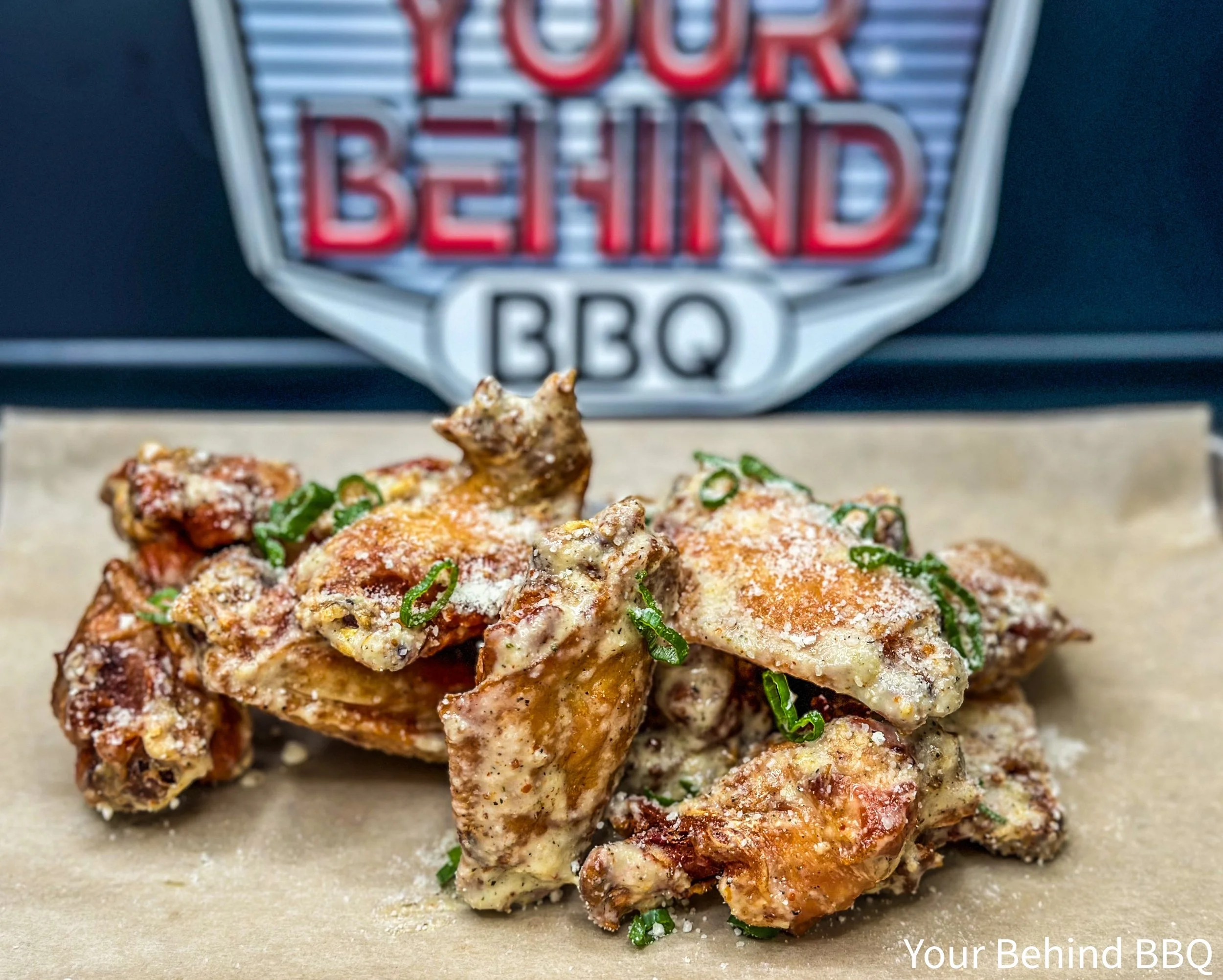 Close-up of grilled barbecued chicken pieces topped with green onions on a sheet of parchment paper, with a blurry sign in the background reading "Your Behind BBQ."