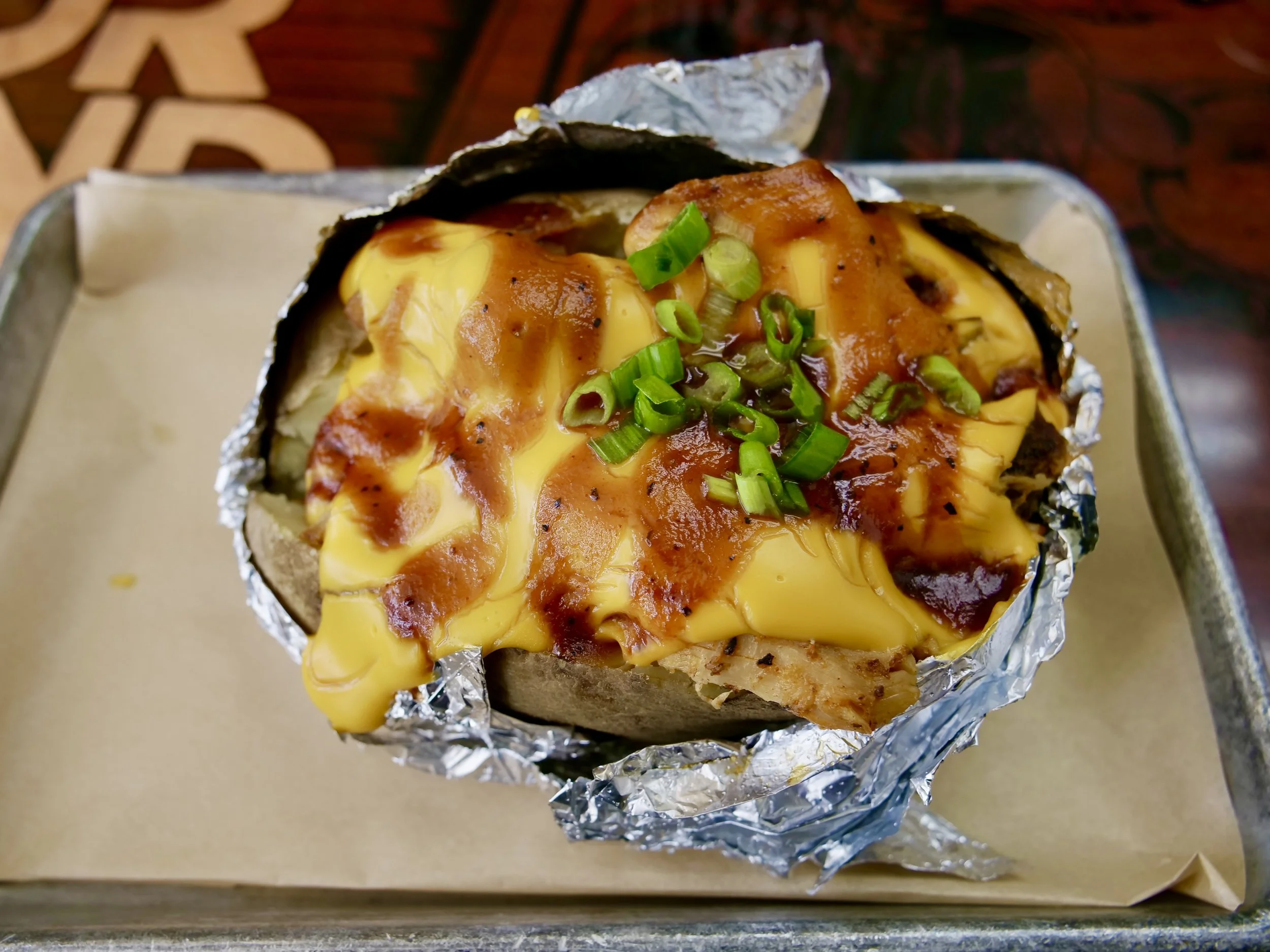Close-up of a baked potato topped with melted cheese, sauce, chopped green onions, wrapped in aluminum foil and placed in a cardboard box.
