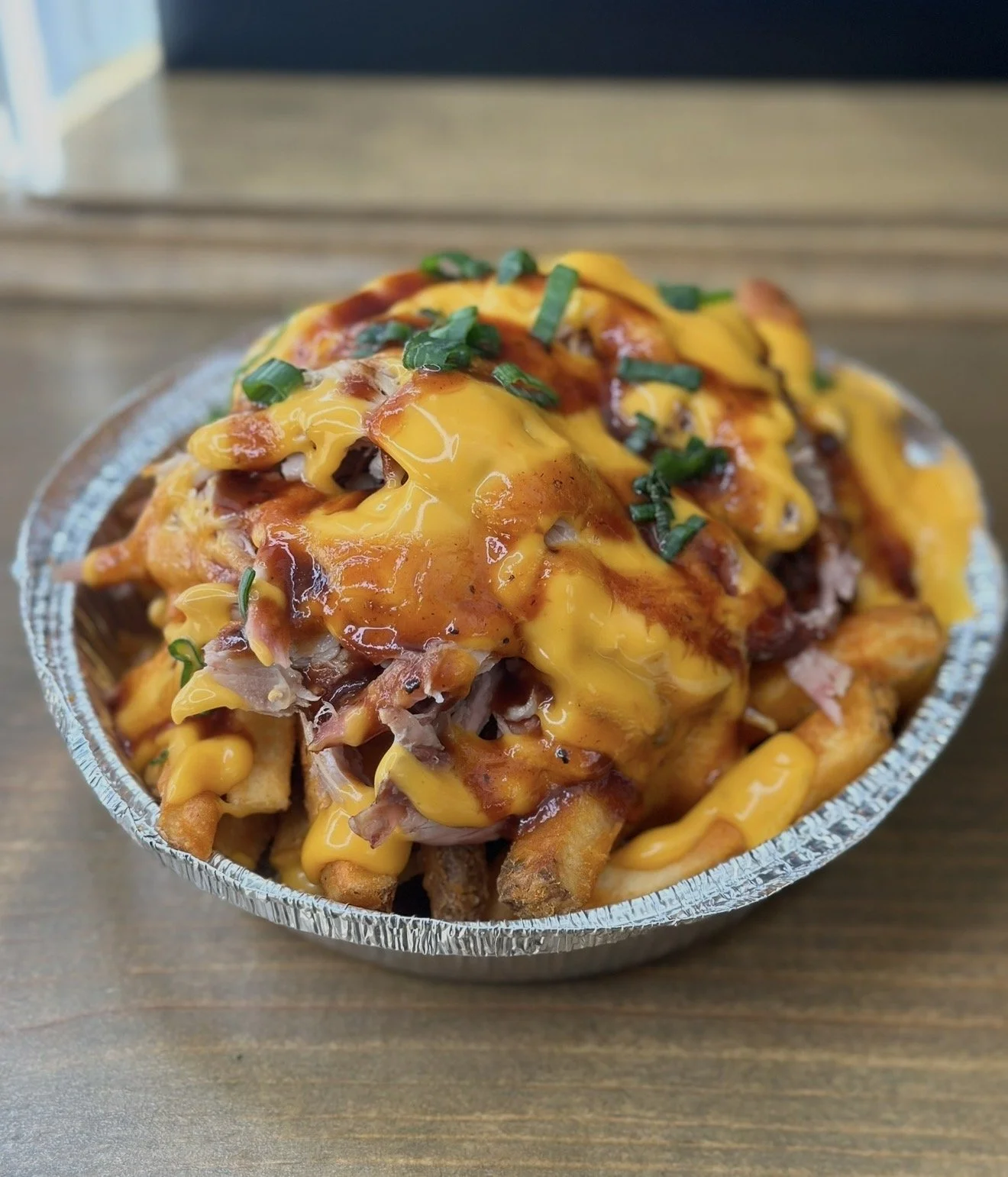 Close-up of a serving of loaded fries topped with melted cheddar cheese, chopped green onions, bacon bits, and barbecue sauce in a foil container on a wooden surface.
