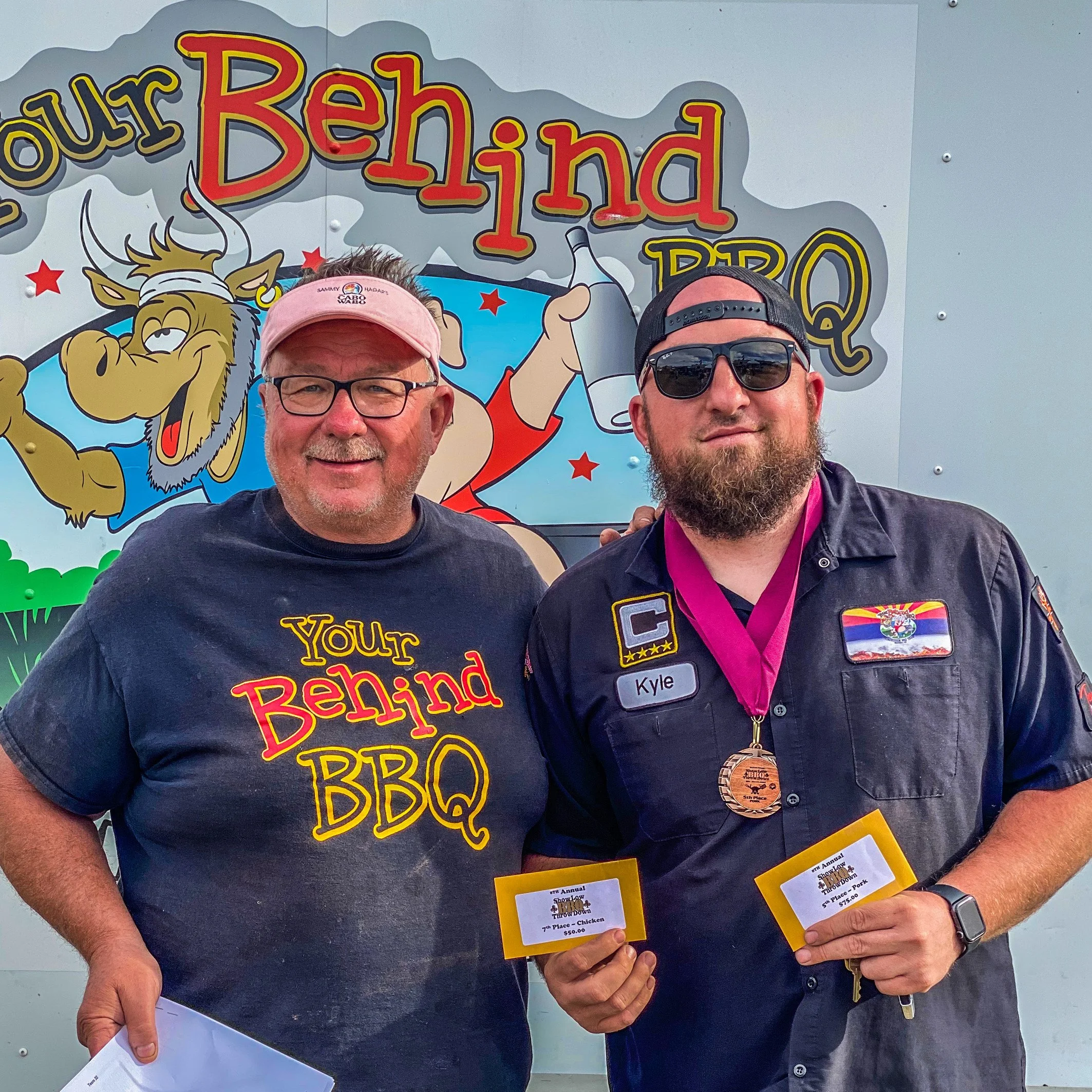 Two smiling men standing in front of a colorful sign with cartoon animal and red stars. One man is wearing a pink baseball cap and glasses, and the other is wearing sunglasses and a black cap. Both are holding yellow and white paper with event details.