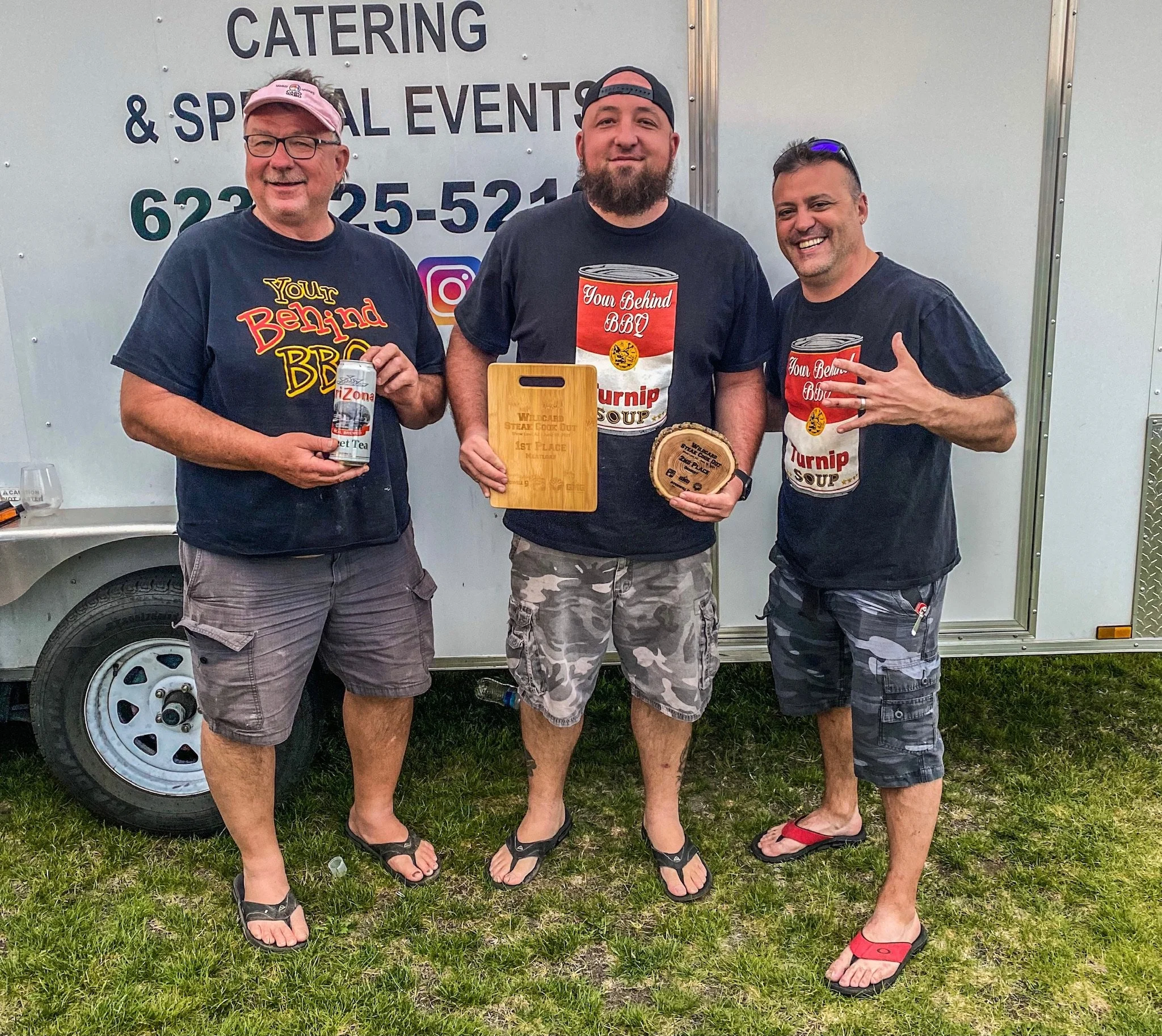 Three men standing outdoors in front of a sign with the text "Catering & Special Events." They are smiling and holding awards, with one holding a can of Arizona tea and another showing a wooden plaque that says "Steak Cook Out 1st Place Meatmaker." All three are wearing black t-shirts and flip-flops.