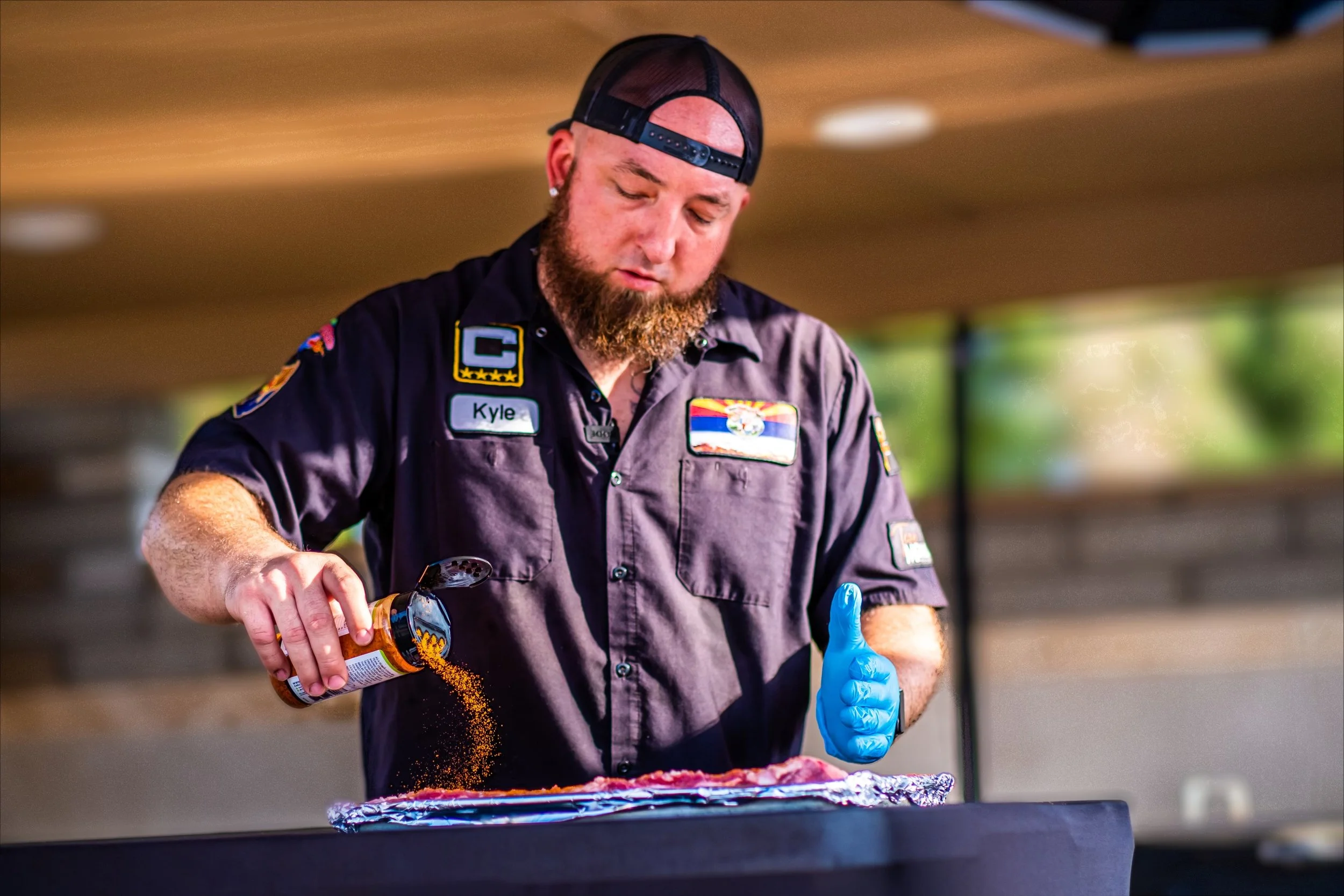 A man with a beard wearing a black cap and dark uniform with patches, pouring seasoning from a bottle onto food while wearing a blue glove.