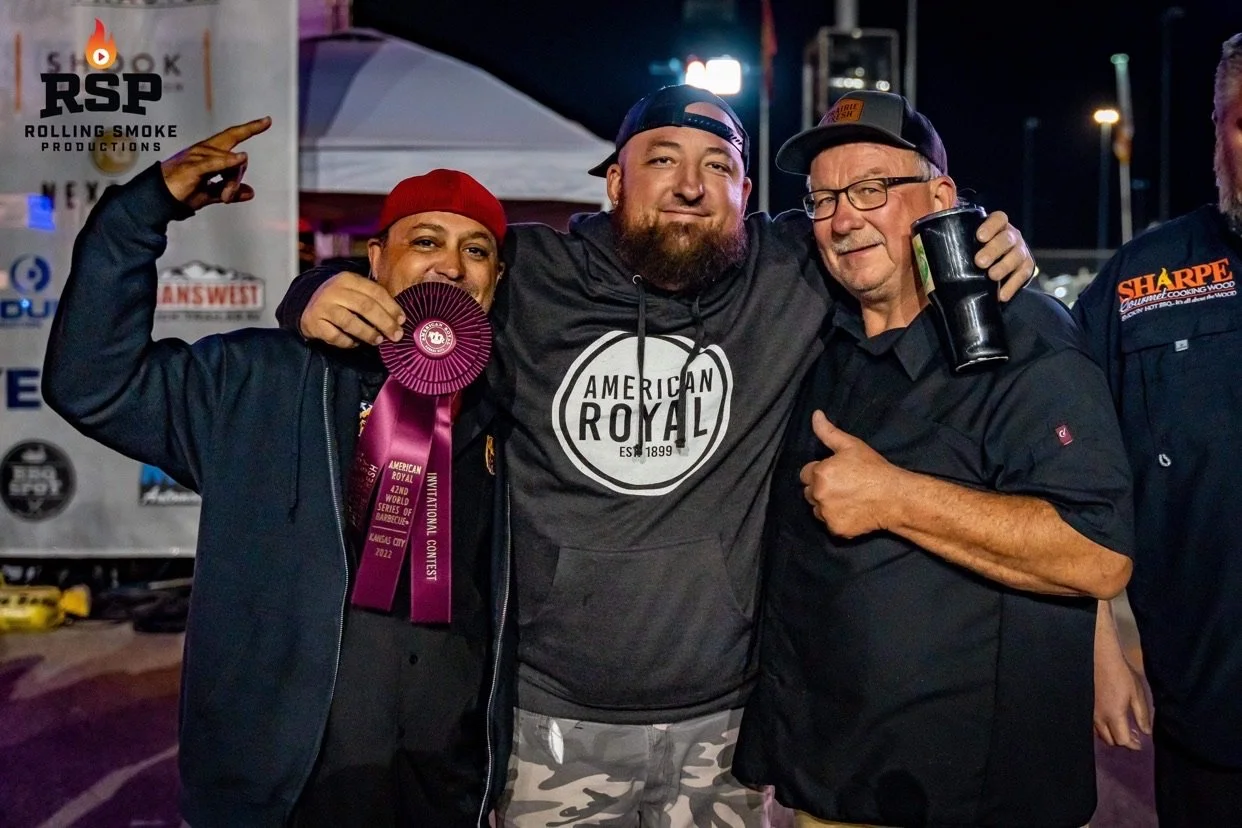 Three men standing together at a nighttime event, smiling and posing for a photo. The man on the left is holding a pink ribbon and ribbon award, the man on the right is holding a drink. There are tents and banners in the background.