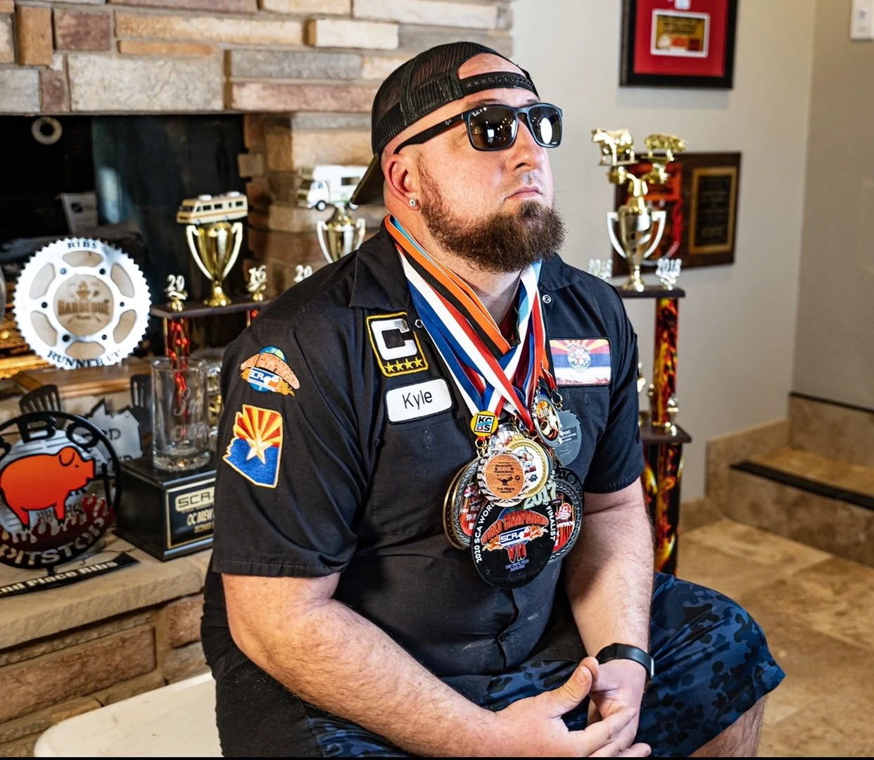 A man with a beard and sunglasses wearing a black shirt with patches and medals around his neck, sitting indoors with a backdrop of trophies and awards.