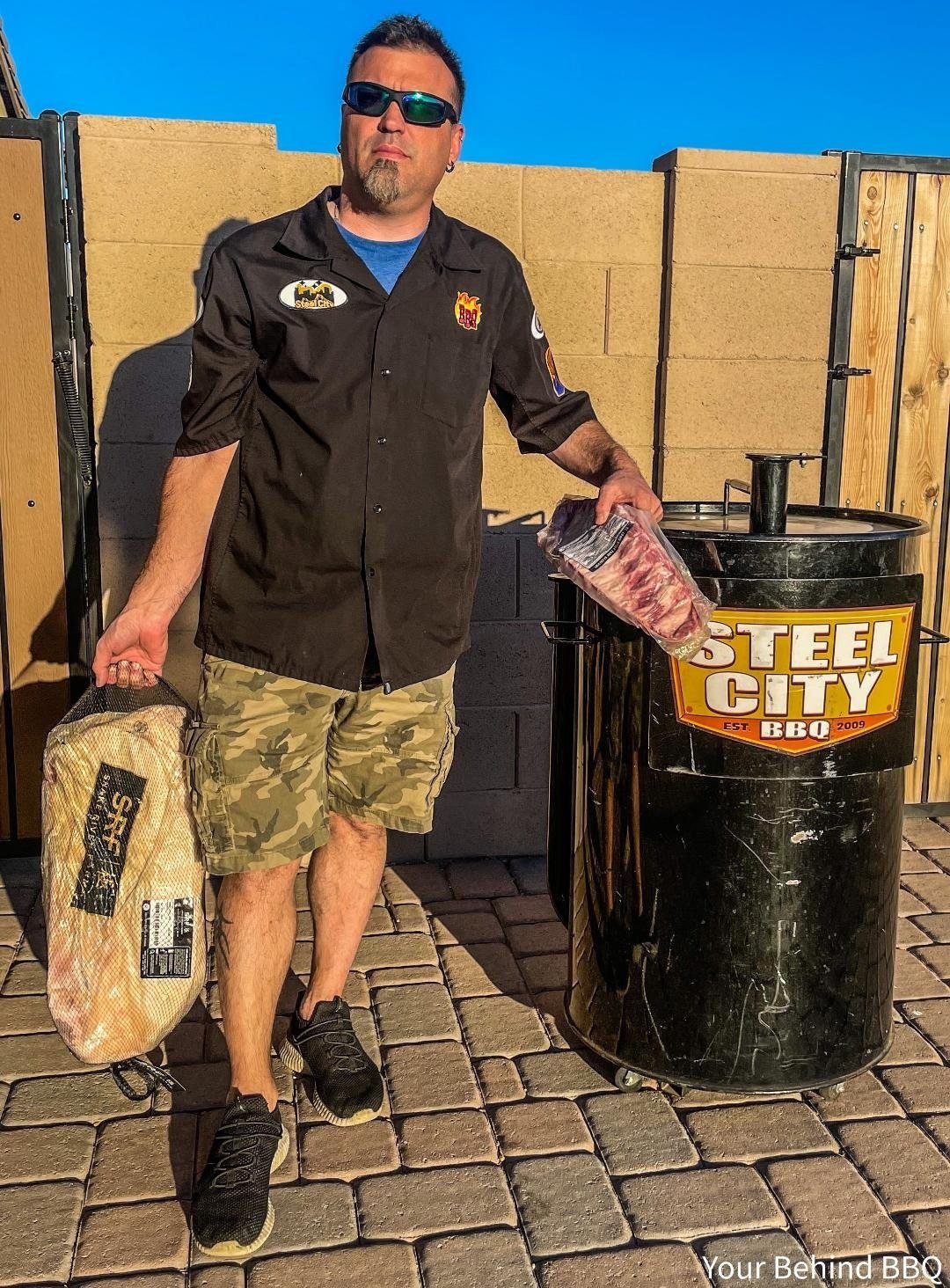 Man wearing sunglasses, black shirt with patches, and camouflage shorts standing outdoors next to a smoker grill with a "Steel City BBQ" logo, holding a package of meat and a bag of wood chips, on a stone-paved patio with a tan brick wall and clear blue sky in the background.