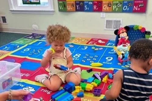Two young children playing with colorful building blocks on a bright educational mat in a classroom.