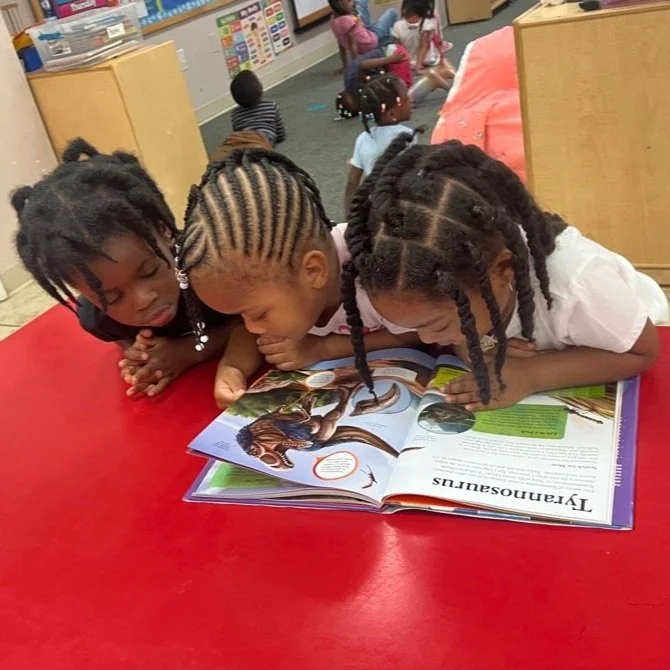 Three young girls with braided hair are reading a picture book together on a red classroom table.