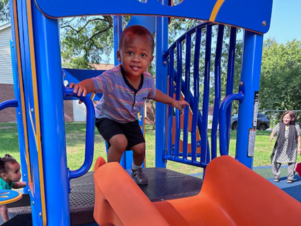 A young boy climbing on a blue and orange playground structure in a park, with a girl standing in the background and another girl sitting nearby. The scene is outdoors with trees and a building visible in the background.