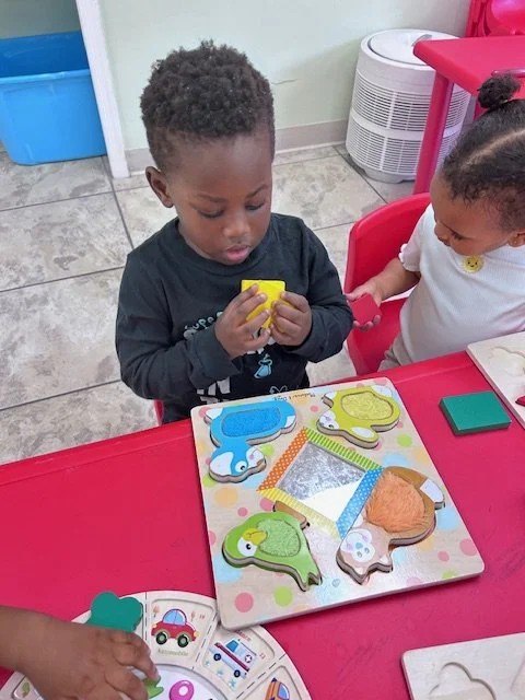 Two young children sitting at a red table, playing with colorful animal-shaped puzzle pieces. The boy on the left, with curly hair, is focusing on a yellow puzzle piece, while the girl on the right, with her hair in a bun, is holding a red puzzle piece. There are various puzzle pieces and a game board on the table.