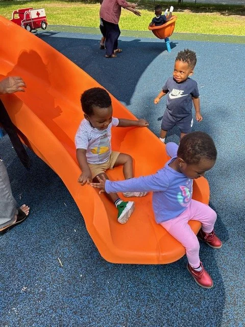 Children playing on an orange slide at a playground, with one girl holding another child's hand at the bottom of the slide and a boy standing nearby with other children and adults in the background.