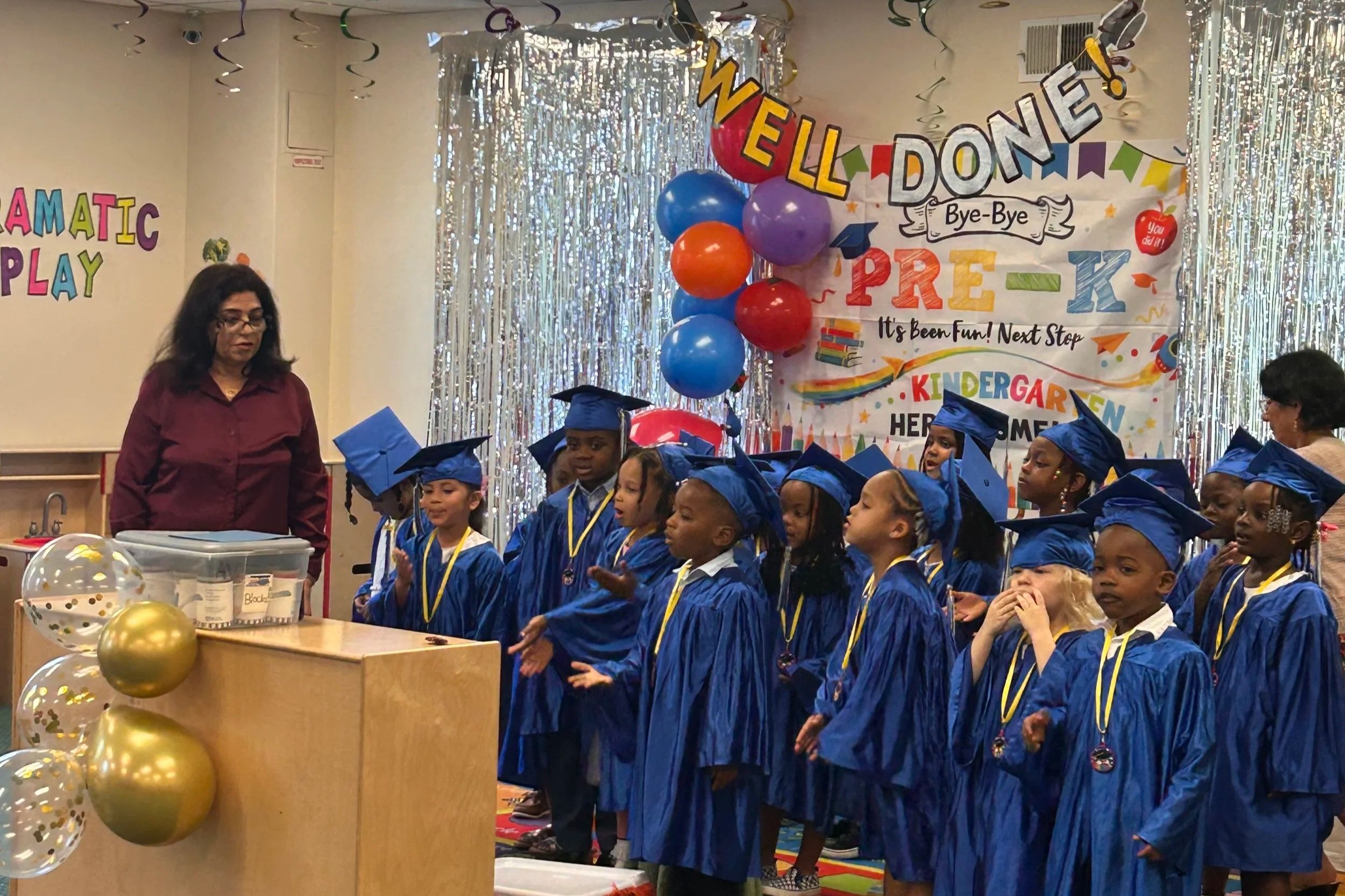 Children in blue graduation gowns and caps standing in a line during a kindergarten graduation ceremony. The background has colorful banners and balloons, and two adults are supervising the event.