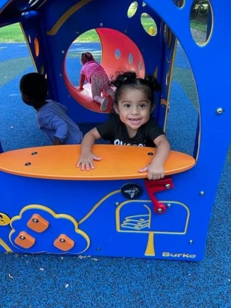 Three young children playing on a colorful playground structure with a blue and orange design. One girl with pigtails is smiling at the camera, while two other children are climbing inside the structure.