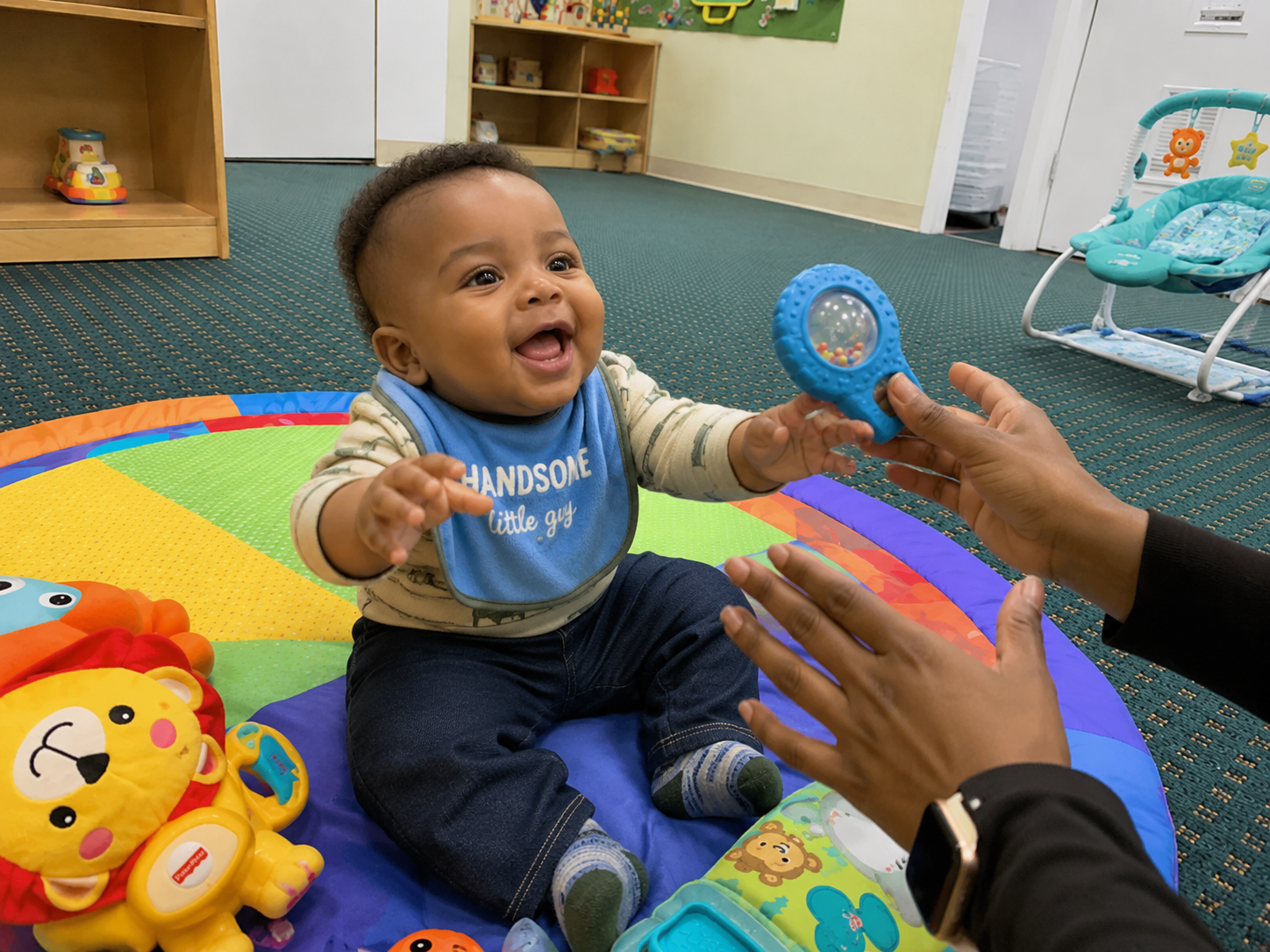 A happy baby sitting on a colorful play mat reaching for a toy handed to him by an adult in a room with toys and a play area.