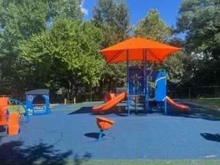 Children's playground with slides, swings, and a large orange shade canopy, located on a blue rubber surface, surrounded by green trees.