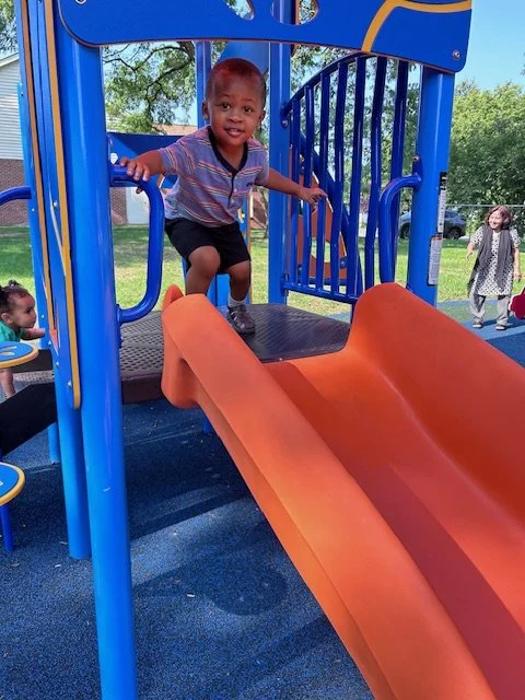 A young child playing on a colorful playground slide outdoors, with other children and trees in the background.