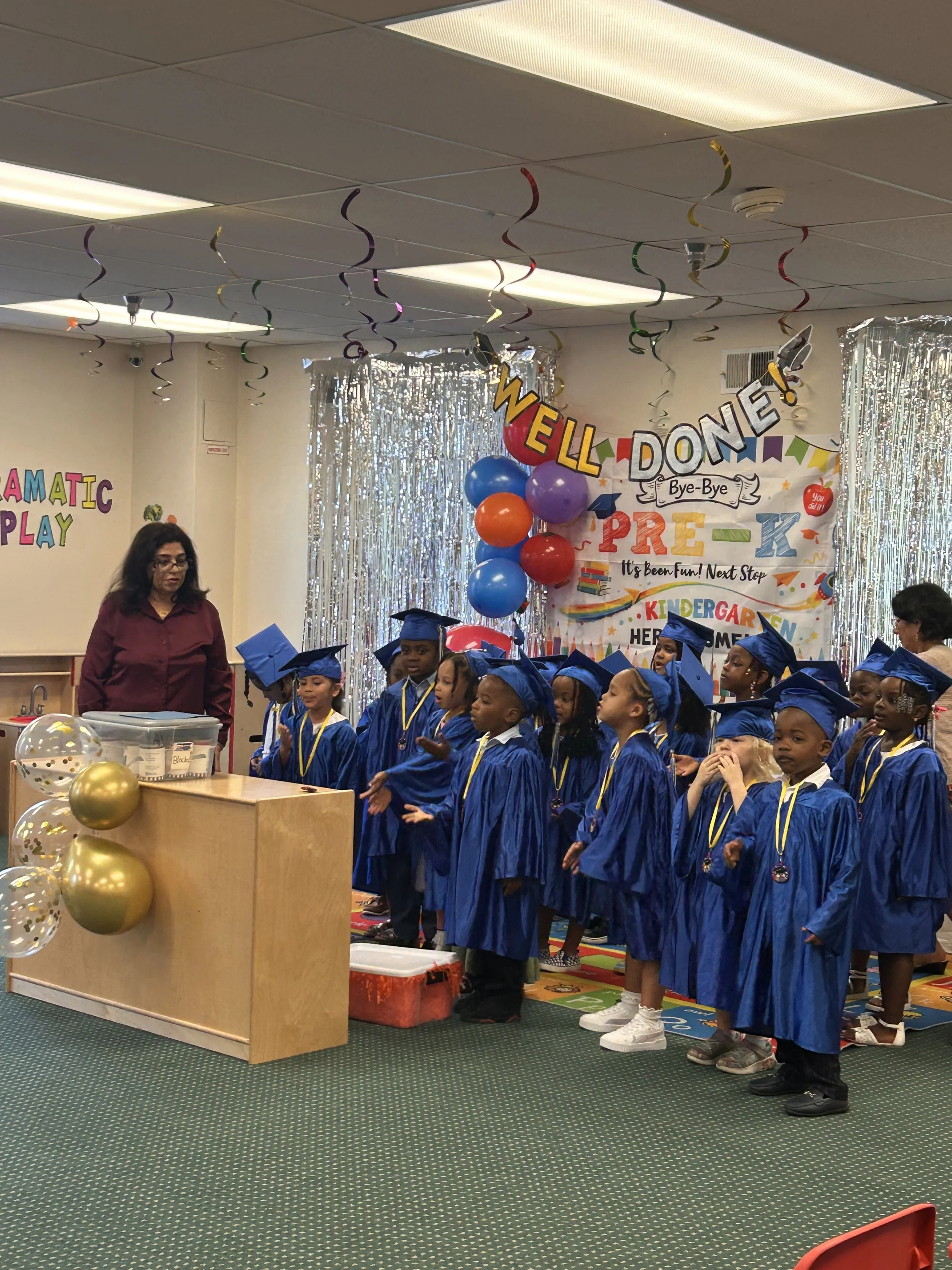 Group of young children in blue graduation gowns and caps standing in a line during a graduation ceremony in a decorated classroom. A woman is standing nearby. The background has shiny silver curtains, colorful balloons, and banners celebrating the event.