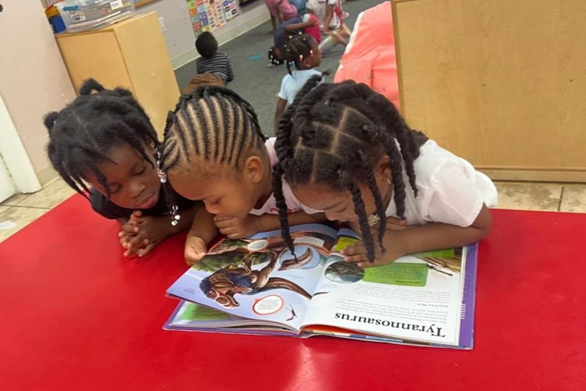 Three young girls with braided hair are lying on a red table, intently looking at a large children's book about dinosaurs. One World Preschool Silver Spring MD best daycares.