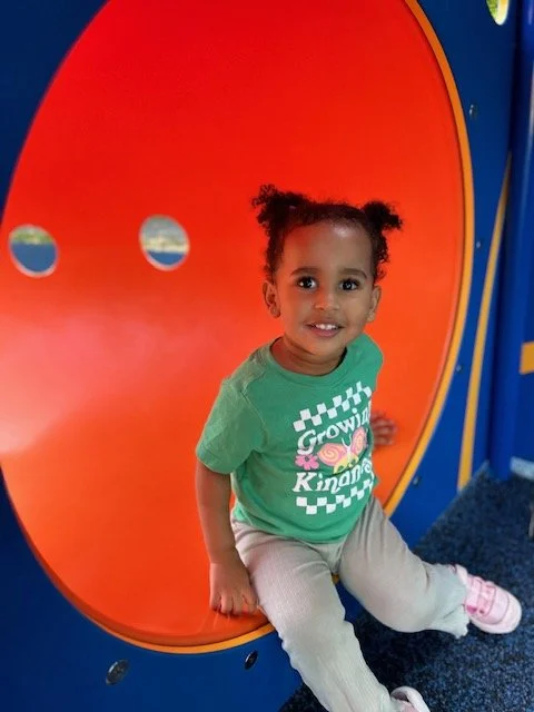 A young girl with curly hair sitting on a colorful playground structure, smiling at the camera.