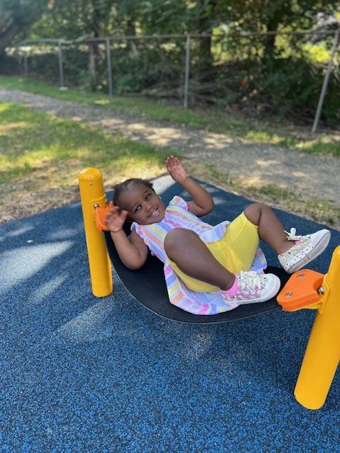 A young girl lying on a black circular swing at a playground, smiling and waving, with trees and a fence in the background.